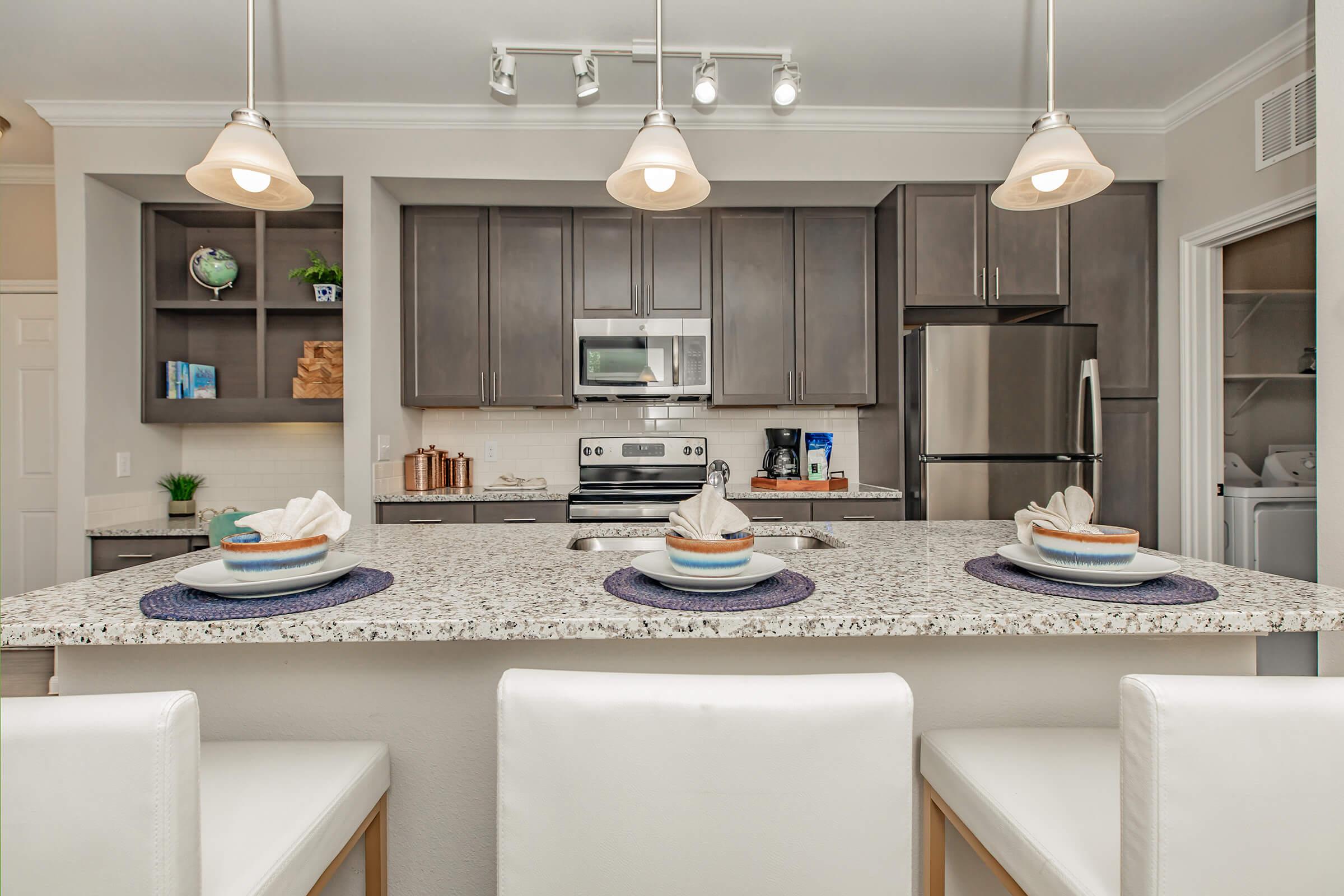 Modern kitchen with dark cabinetry and stainless steel appliances. A large granite island with three plates and napkins set for dining is in the foreground. Bright pendant lights hang above, illuminating the space. Shelves hold decorative items, and a laundry area can be seen in an adjacent room.