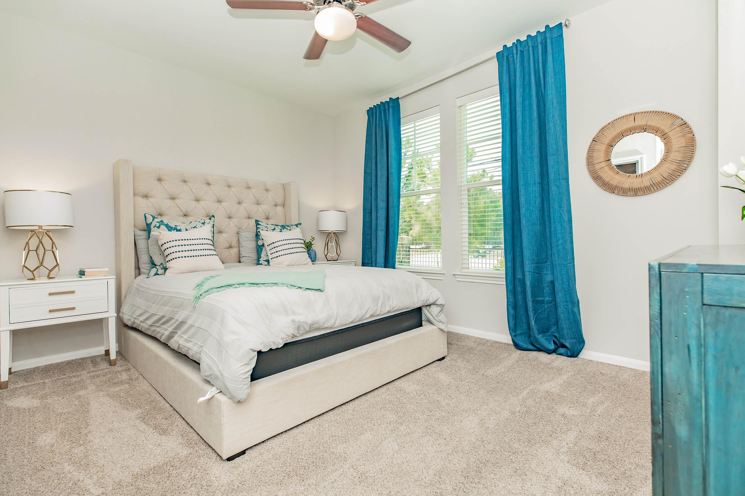 A well-decorated bedroom featuring a beige upholstered bed with decorative pillows, a nightstand with a lamp, and a teal accent decor. Large windows with blue curtains allow natural light in, and a round mirror hangs on the wall. The room has light-colored walls and soft, plush carpeting.