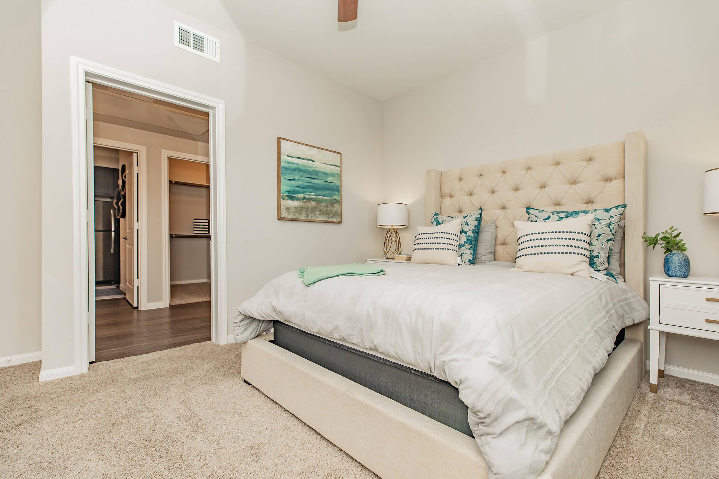 A cozy bedroom featuring a plush bed with decorative pillows, a soft gray blanket, and a neutral-colored headboard. A bedside table with a lamp and a small plant is beside the bed. There's an open door leading to a closet space. The walls are painted light-colored, creating a warm and inviting atmosphere.