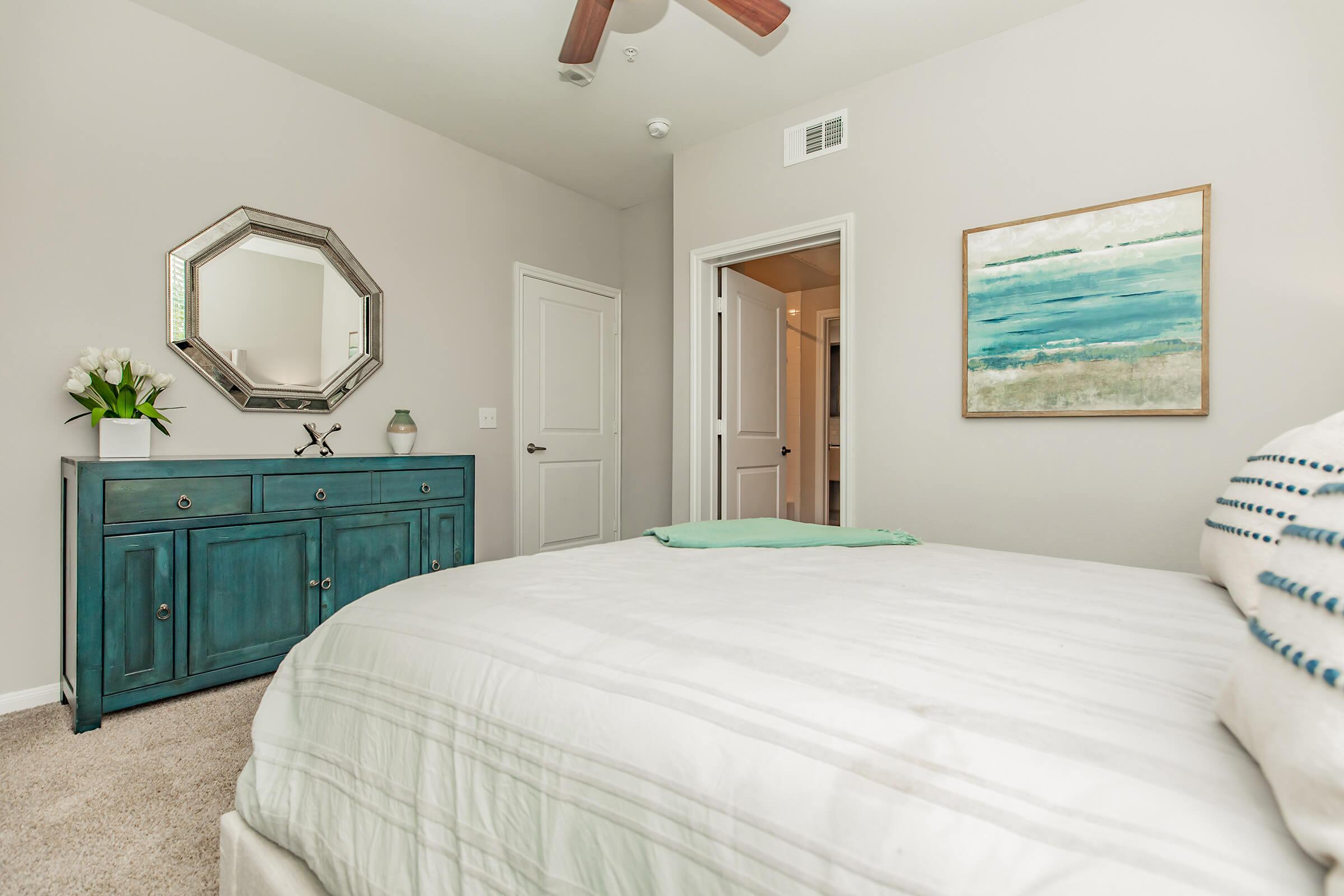 A cozy bedroom featuring a bed with a light-colored striped duvet, a teal dresser, and an abstract painting with blue hues on the wall. The room has a ceiling fan and a mirror reflecting natural light. A door leads to another space, adding to the inviting atmosphere.