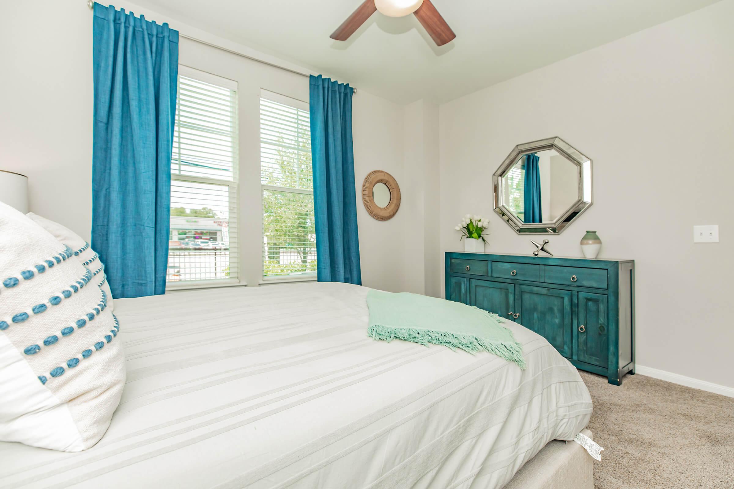 A cozy bedroom featuring a light gray bed with decorative pillows, a turquoise throw blanket, and large windows dressed in blue curtains. A stylish mirror hangs on the wall, and a teal dresser with decorative accents is visible. The room has neutral tones and a soft carpet, creating a welcoming atmosphere.