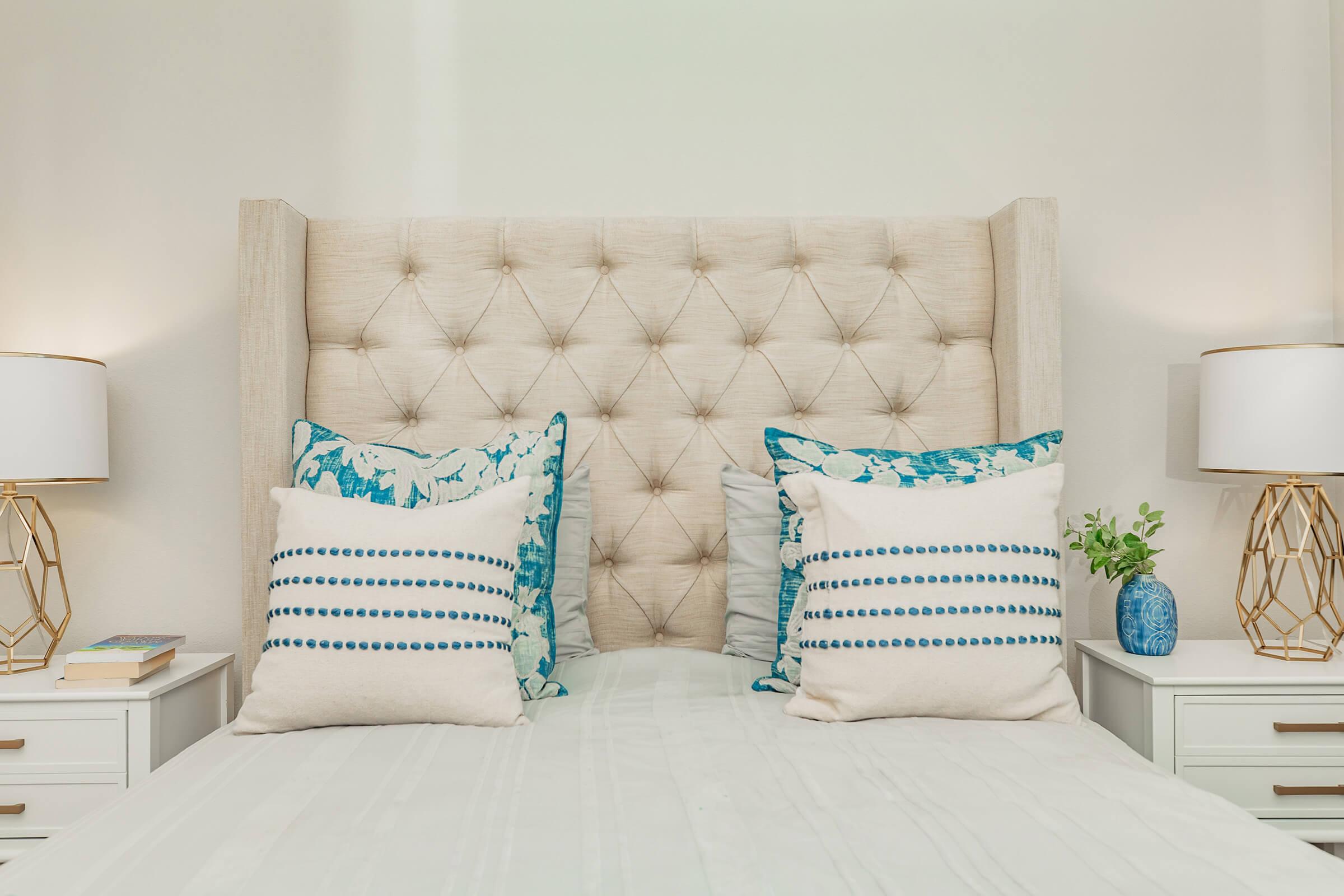 A cozy bedroom scene featuring a tufted beige headboard with decorative pillows in blue and white patterns. Two stylish bedside lamps with geometric bases flank the bed, which is made with white linens. A small plant and a book add a touch of decor on the nightstand.