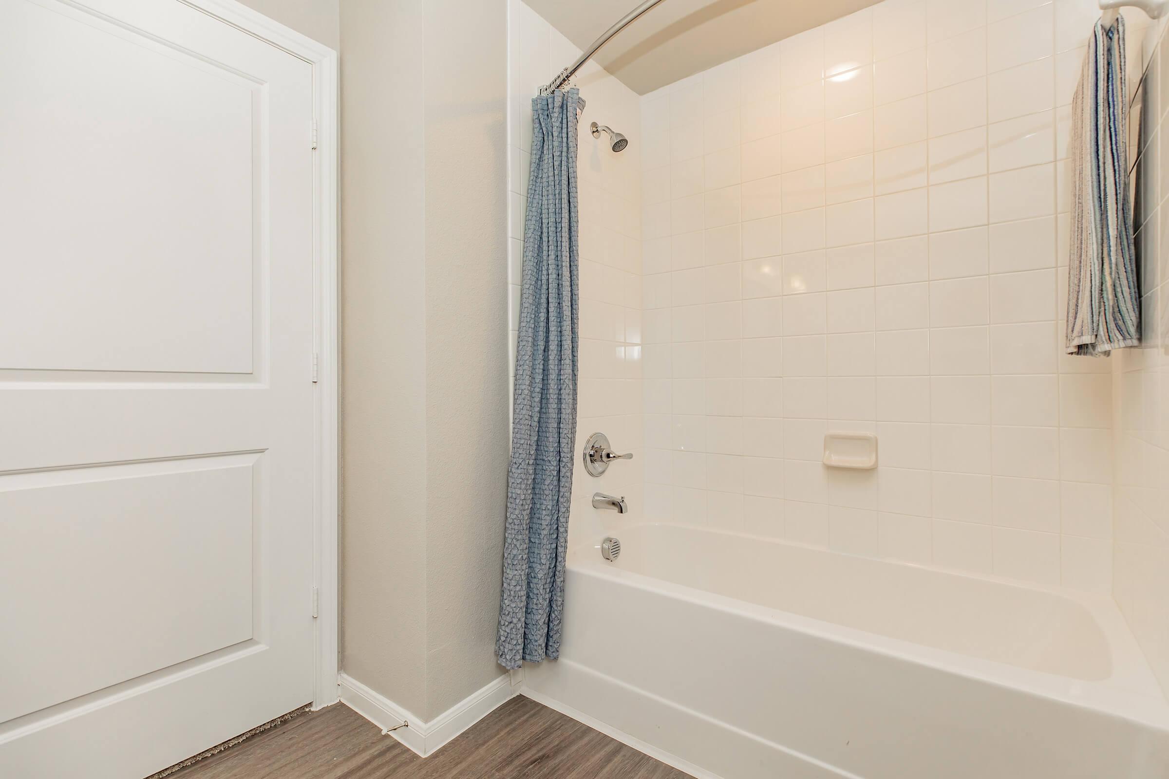 A clean bathroom featuring a white bathtub with a curved shower rod, light blue shower curtain, and tiled walls. There is a towel hanging nearby, and a closed white door leads out of the bathroom. The floor is made of light-colored wood.