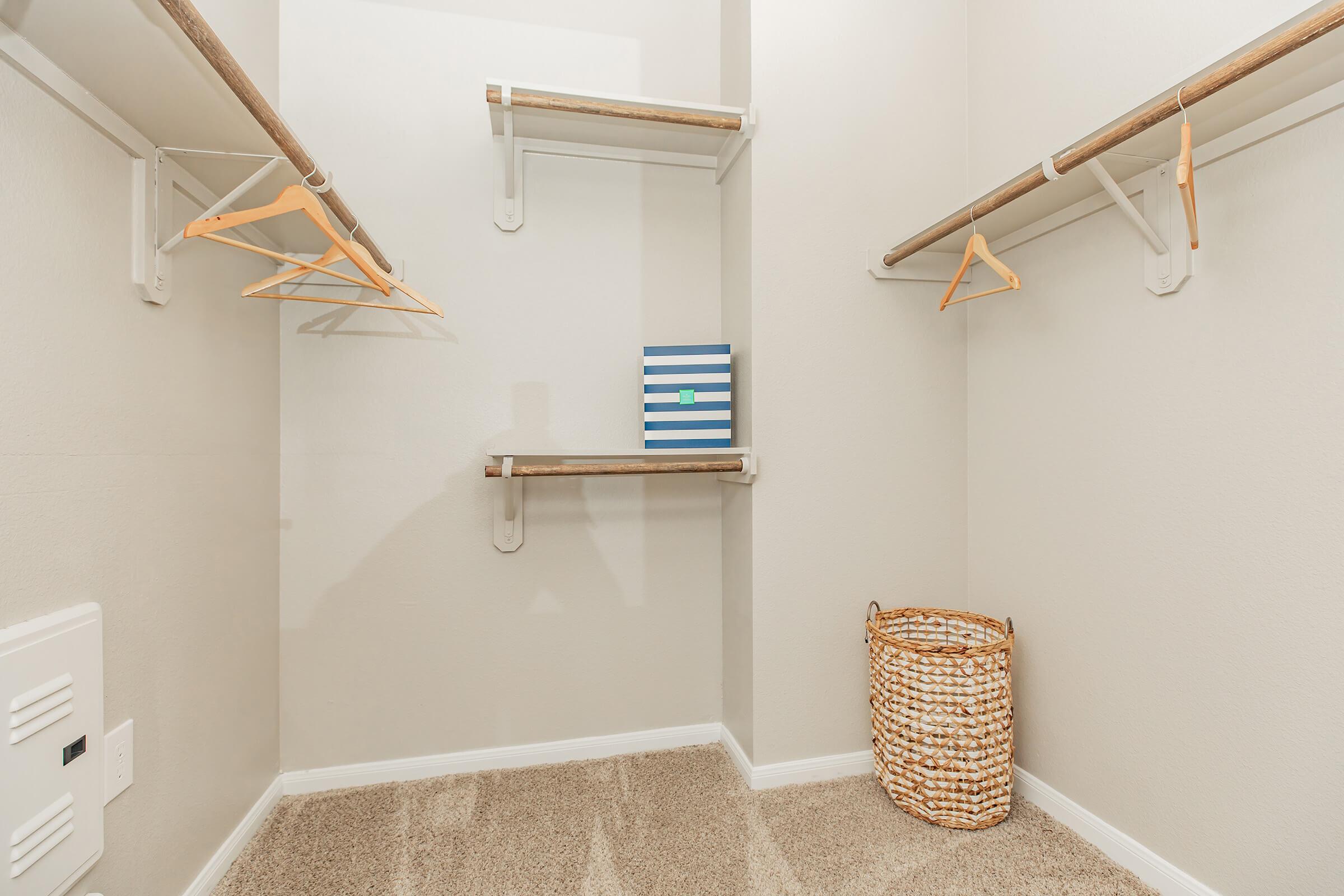 Empty closet with light gray walls, featuring two wooden hanging rods with orange hangers. There are three shelves attached to the walls, and a striped blue and white box is placed on one of the shelves. A woven basket sits in the corner on a beige carpeted floor.
