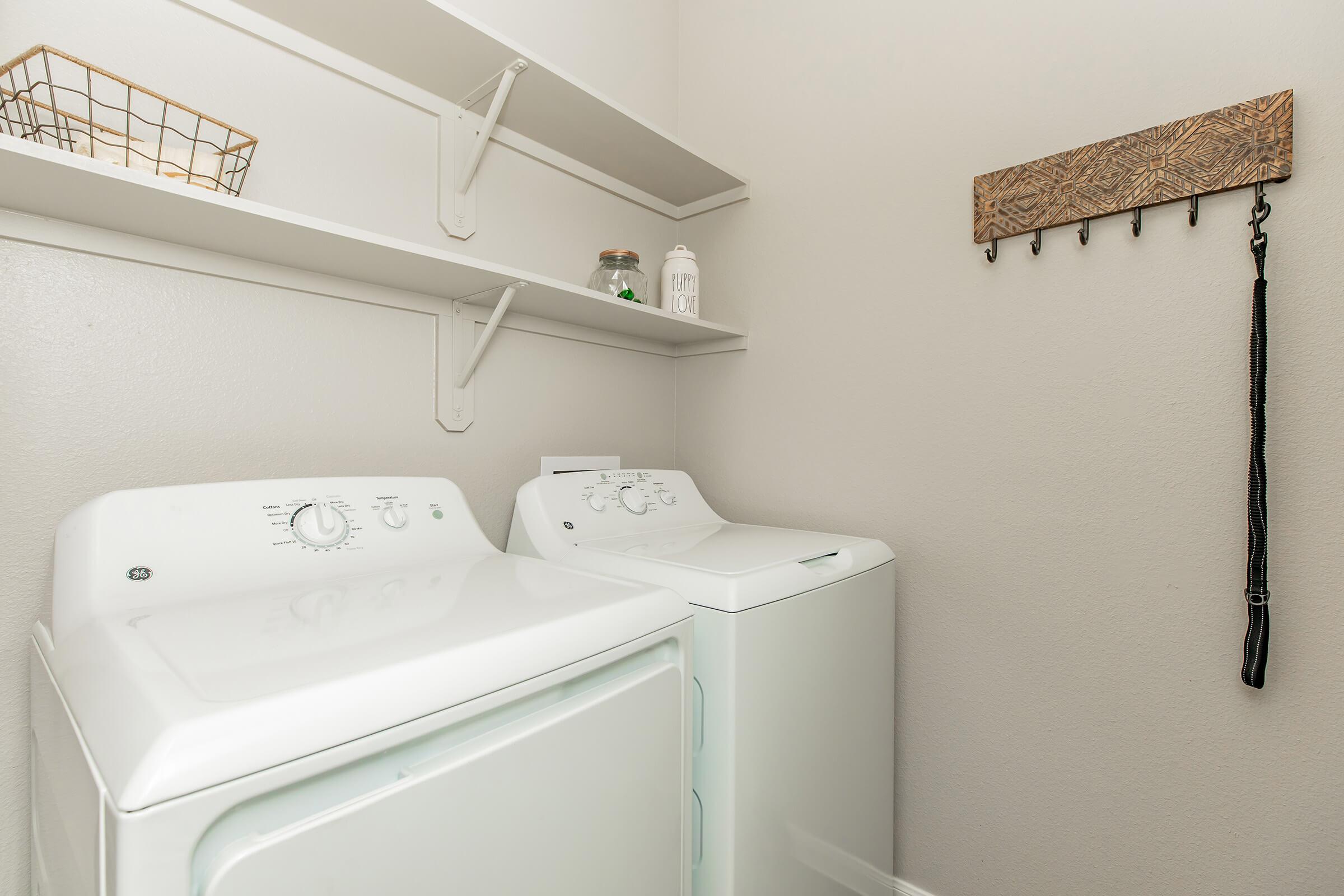 A clean laundry room featuring a white washing machine and dryer side by side. Above them, there are simple white shelves holding a jar and a decorative lantern. A wooden wall hook with multiple hooks is mounted beside the shelves, enhancing the functional and organized look of the space.
