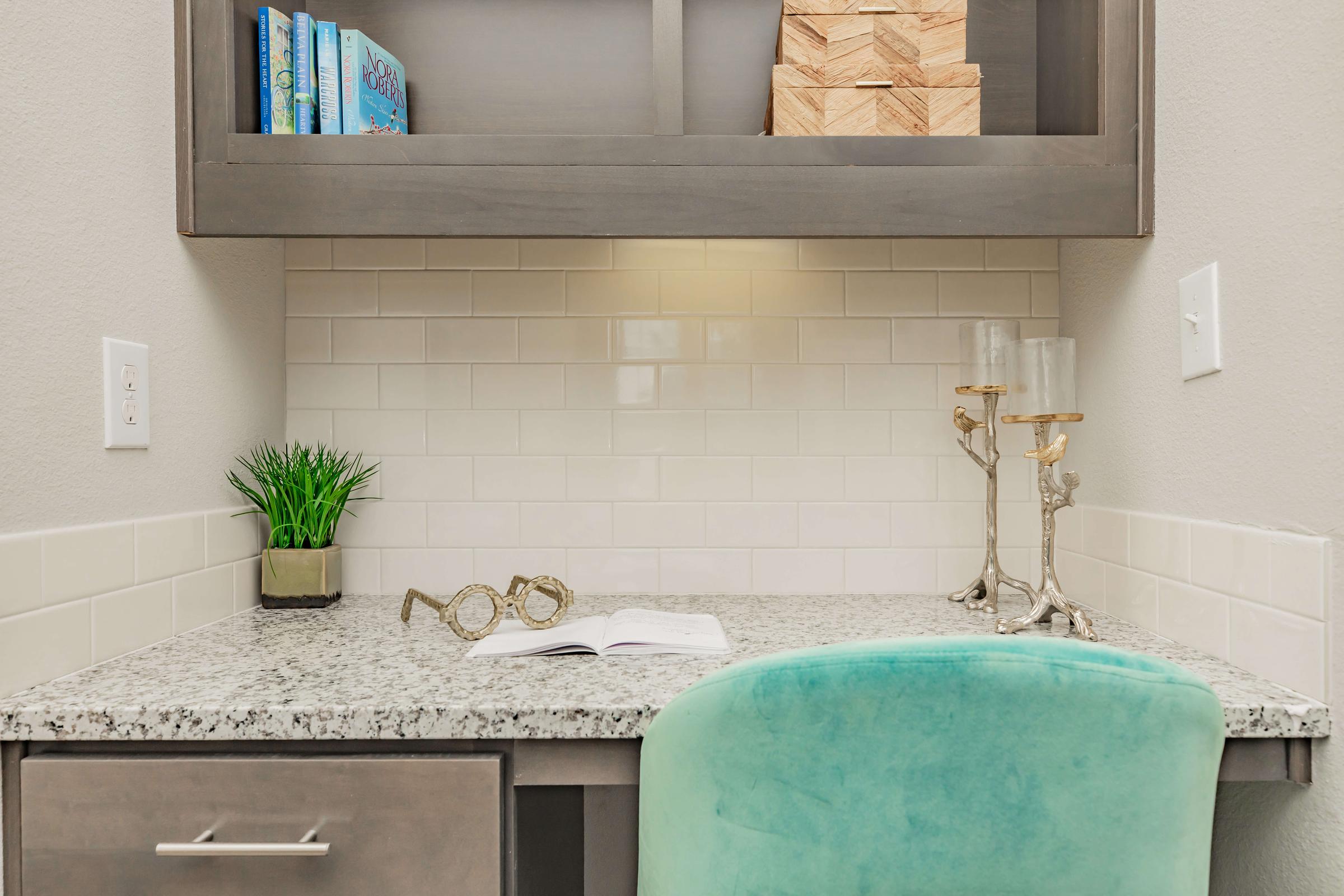 A neatly organized workspace featuring a light gray desk with a granite surface, a teal chair, and a small plant. Above the desk, there are shelves holding blue books and wooden boxes. Two decorative candle holders sit alongside an open book, creating an inviting study area.