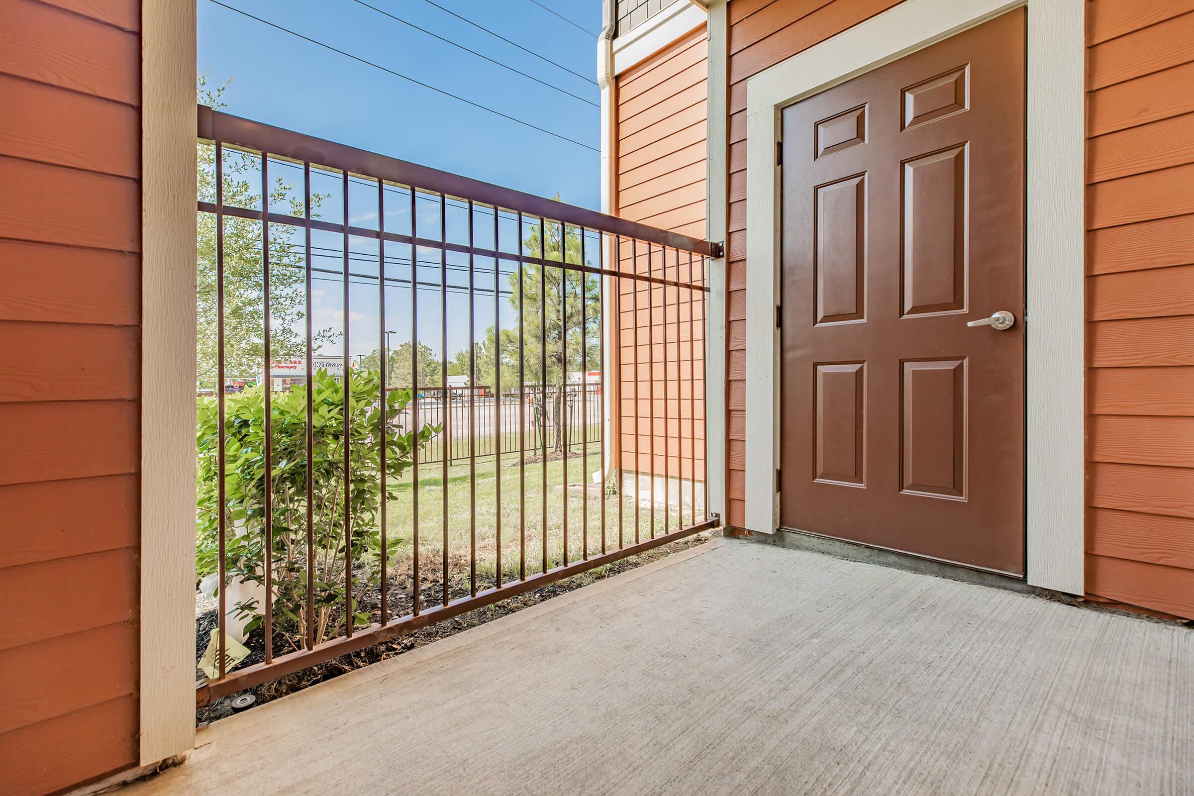 A view of a patio area outside a brown door, with orange wooden siding on the walls and a metal railing. The floor is concrete, and there is a small patch of grass and shrubs visible beyond the railing, under a clear blue sky.