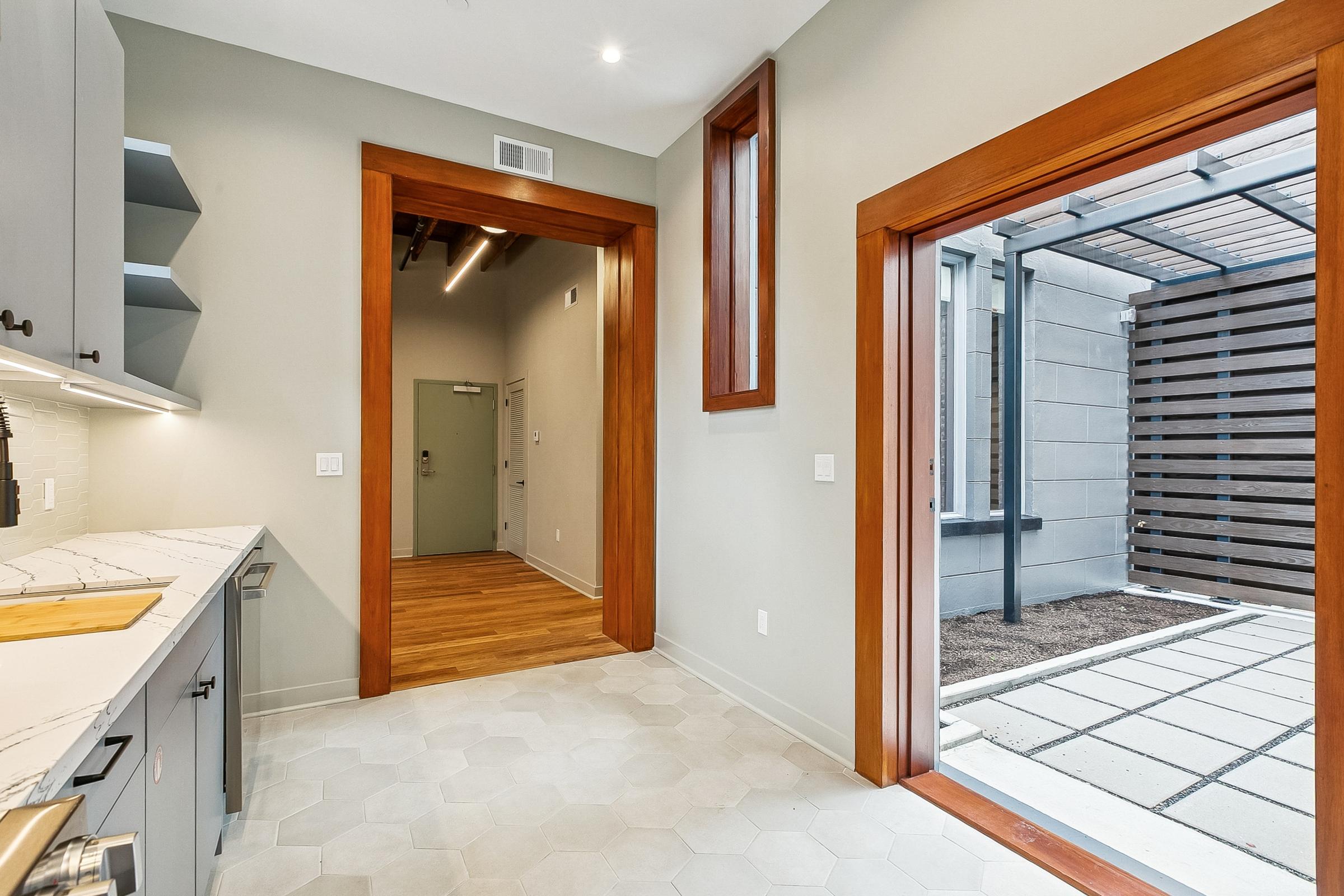 Interior view of a modern kitchen and hallway featuring light gray cabinetry, a marble countertop, and hexagonal tile flooring. A doorway leads to an outdoor area with a patio and wooden privacy slats. Natural light fills the space through large windows and doors.