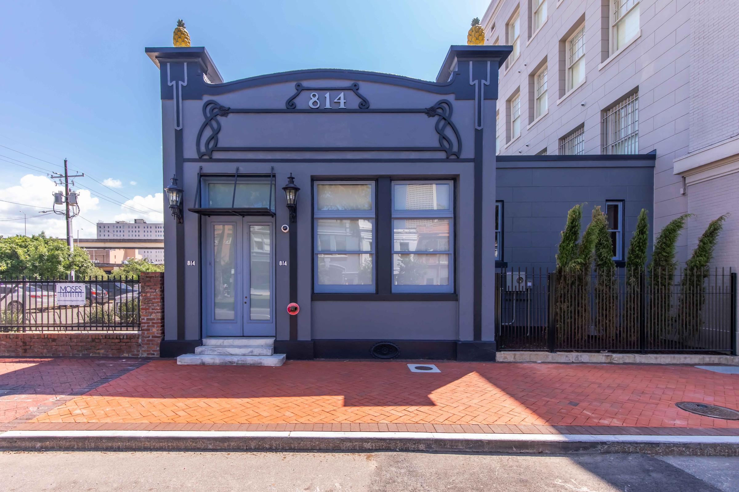 A blue-gray single-story building with decorative details, featuring a white door and windows. The address "814" is prominently displayed above the door. The facade has black accents and two small pineapples atop the roof. The surrounding area includes brick pavement and low shrubs, set against a clear blue sky.