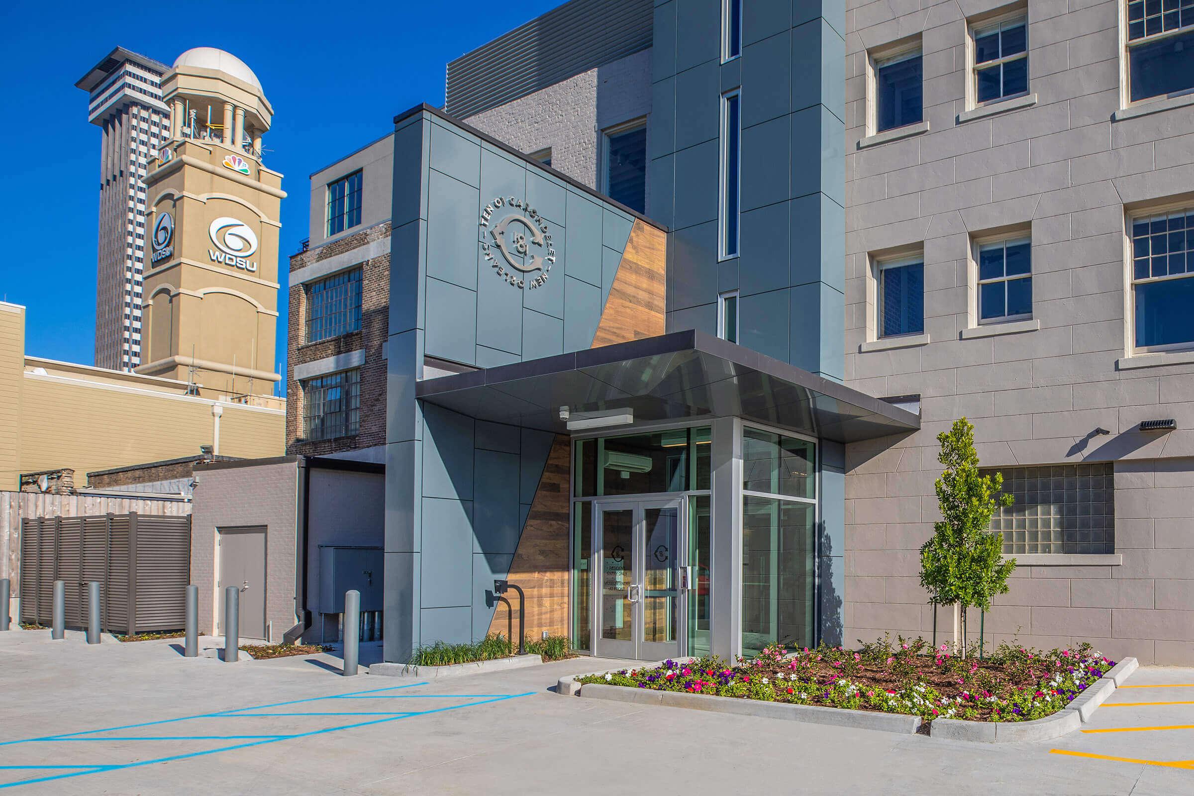 Modern building entrance featuring a glass door, decorative wood and stone accents, and landscaped flower beds. In the background, a tall clock tower with a logo is visible against a clear blue sky. The area is clean and well-maintained, with parking spaces lined out in front.