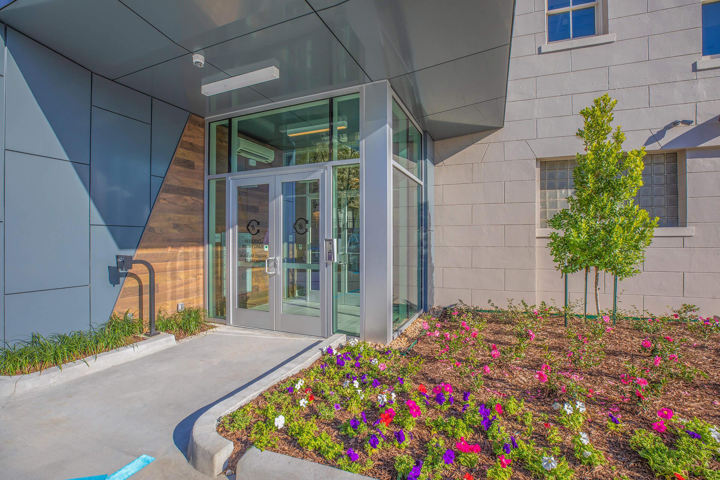 Modern entrance of a building featuring large glass doors, surrounded by colorful flower beds with various blooms. The facade includes a combination of stone and metal materials, with a wooden accent wall visible to the left. Lush greenery adds to the inviting atmosphere.