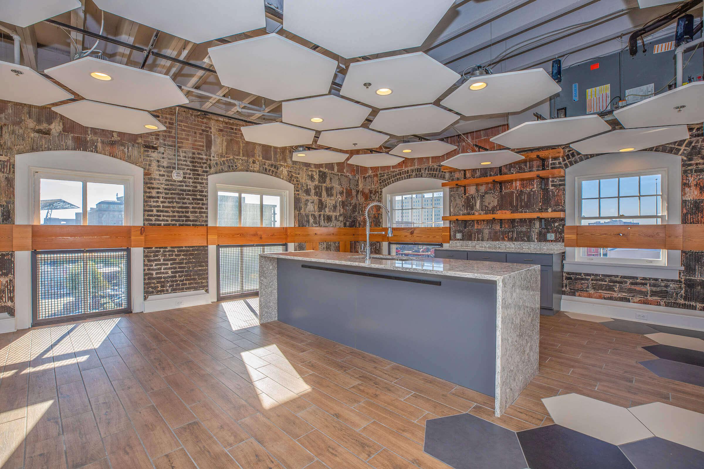 Modern kitchen interior featuring a central island with a sleek gray countertop, surrounded by hexagonal ceiling panels and exposed brick walls. The space has large windows that provide natural light, wooden shelves on the sides, and a contemporary design that blends industrial and modern aesthetics.