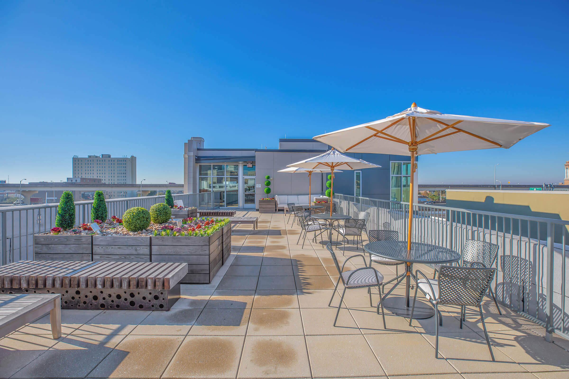 A rooftop terrace featuring lounge chairs, dining tables with umbrellas, and planters with greenery and flowers. The background shows a clear blue sky and distant buildings, creating a bright and inviting outdoor space for relaxation and gatherings.