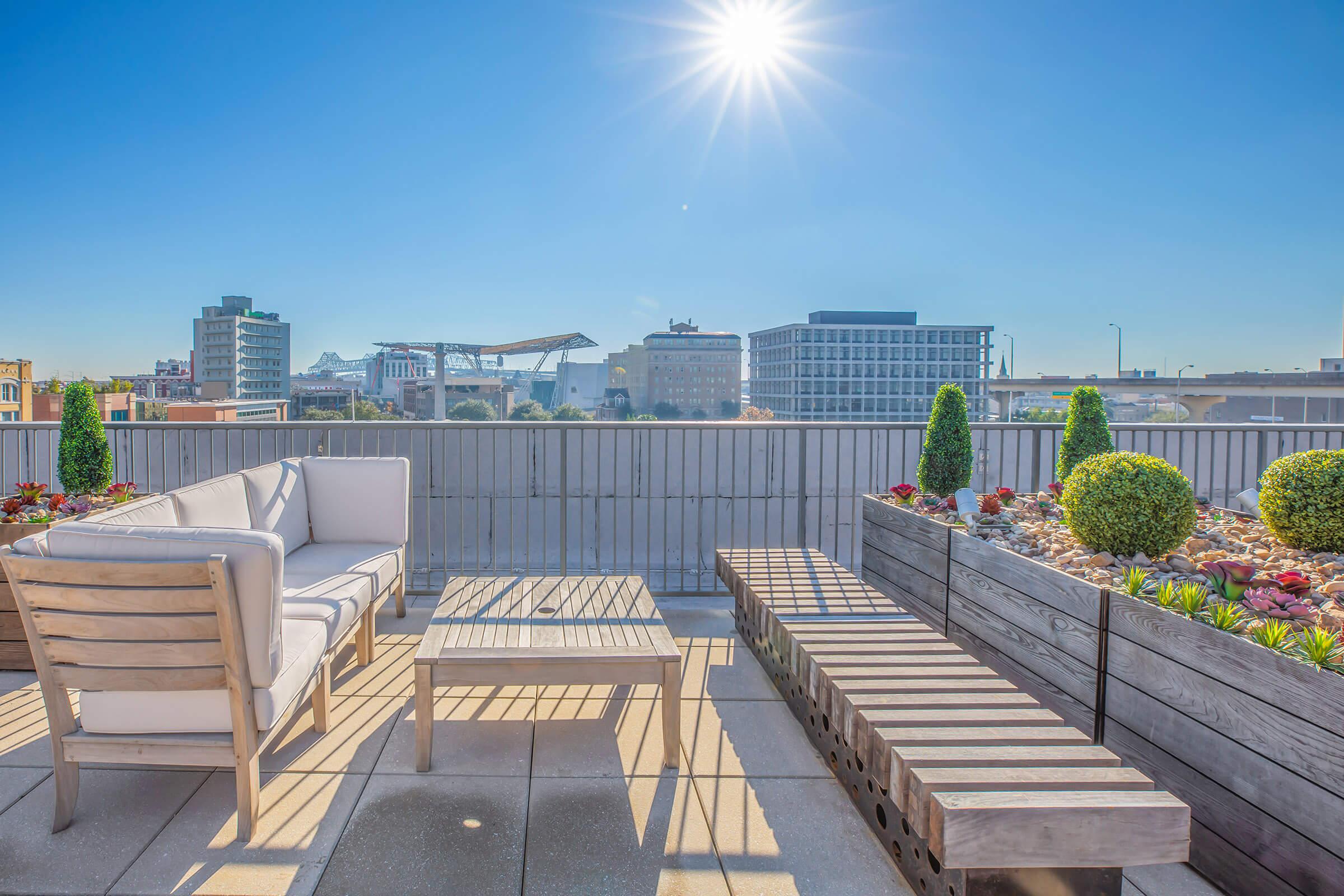 A modern rooftop terrace featuring comfortable outdoor furniture, including a couch and coffee table, surrounded by landscaped planters and succulents. The skyline in the background includes various buildings and a bright sun shining in a clear blue sky.