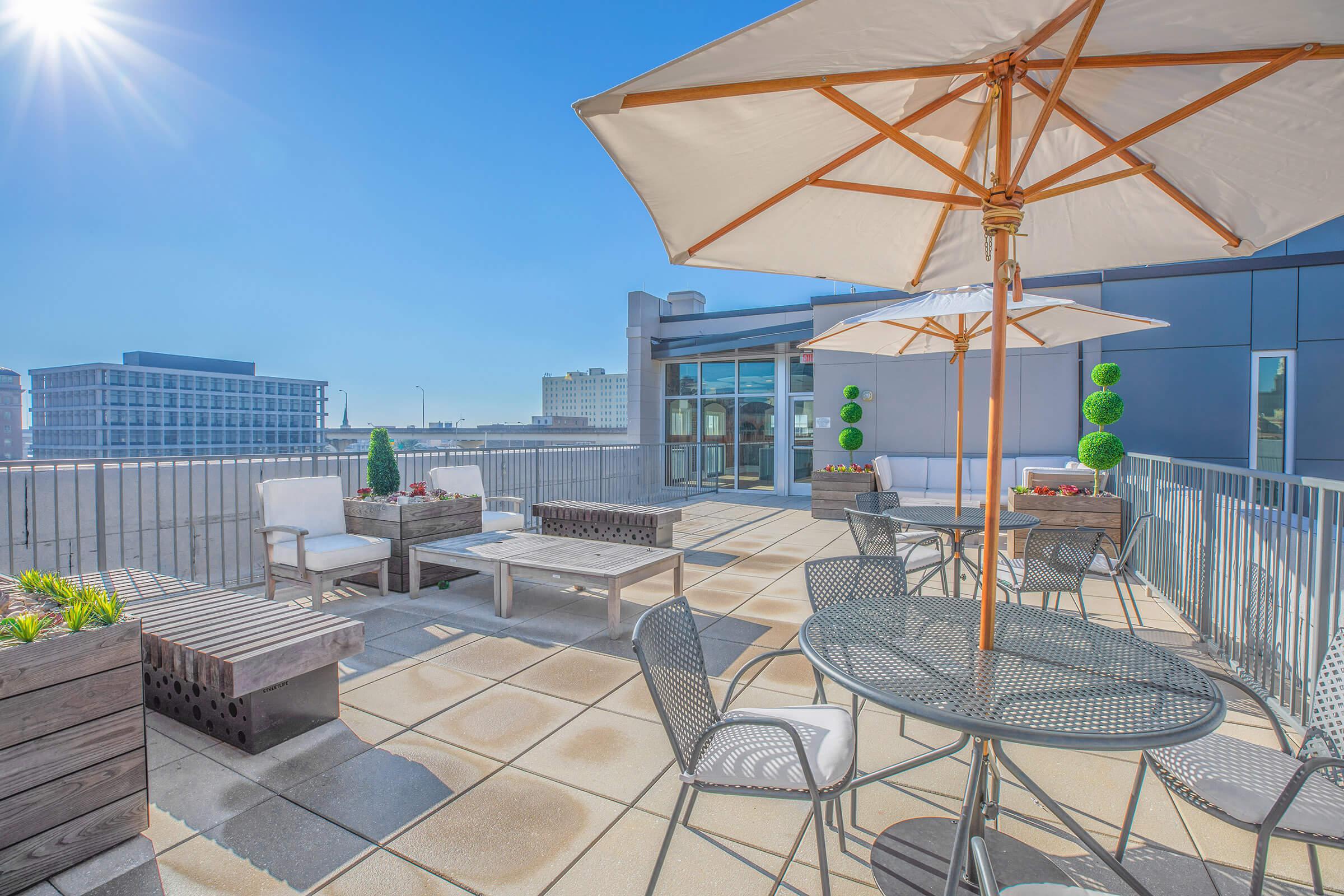 A sunny rooftop terrace featuring several seating areas, including a round table with chairs, lounge seating, and decorative plants. An umbrella provides shade, and there are modern buildings visible in the background against a clear blue sky.