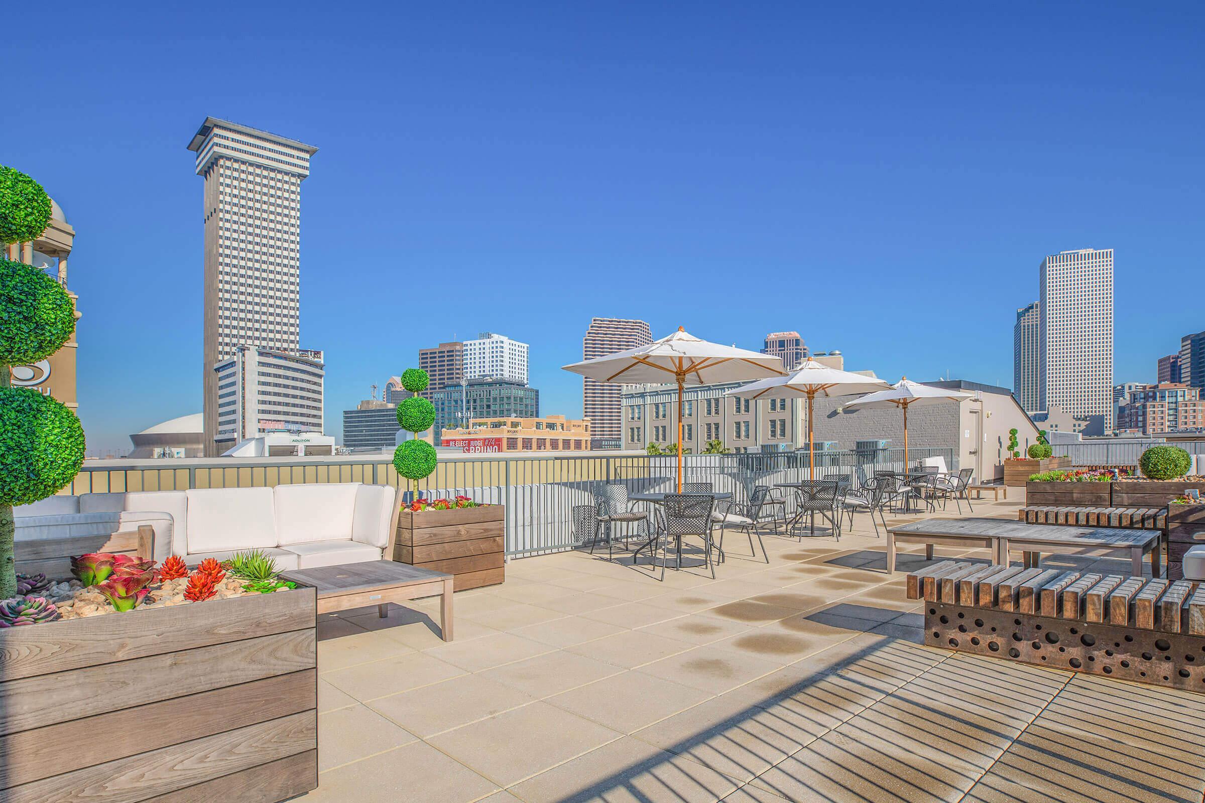 Rooftop terrace featuring comfortable seating, potted plants, and tables under umbrellas. The city skyline with modern buildings is visible in the background against a clear blue sky. The space is designed for relaxation and outdoor gatherings.