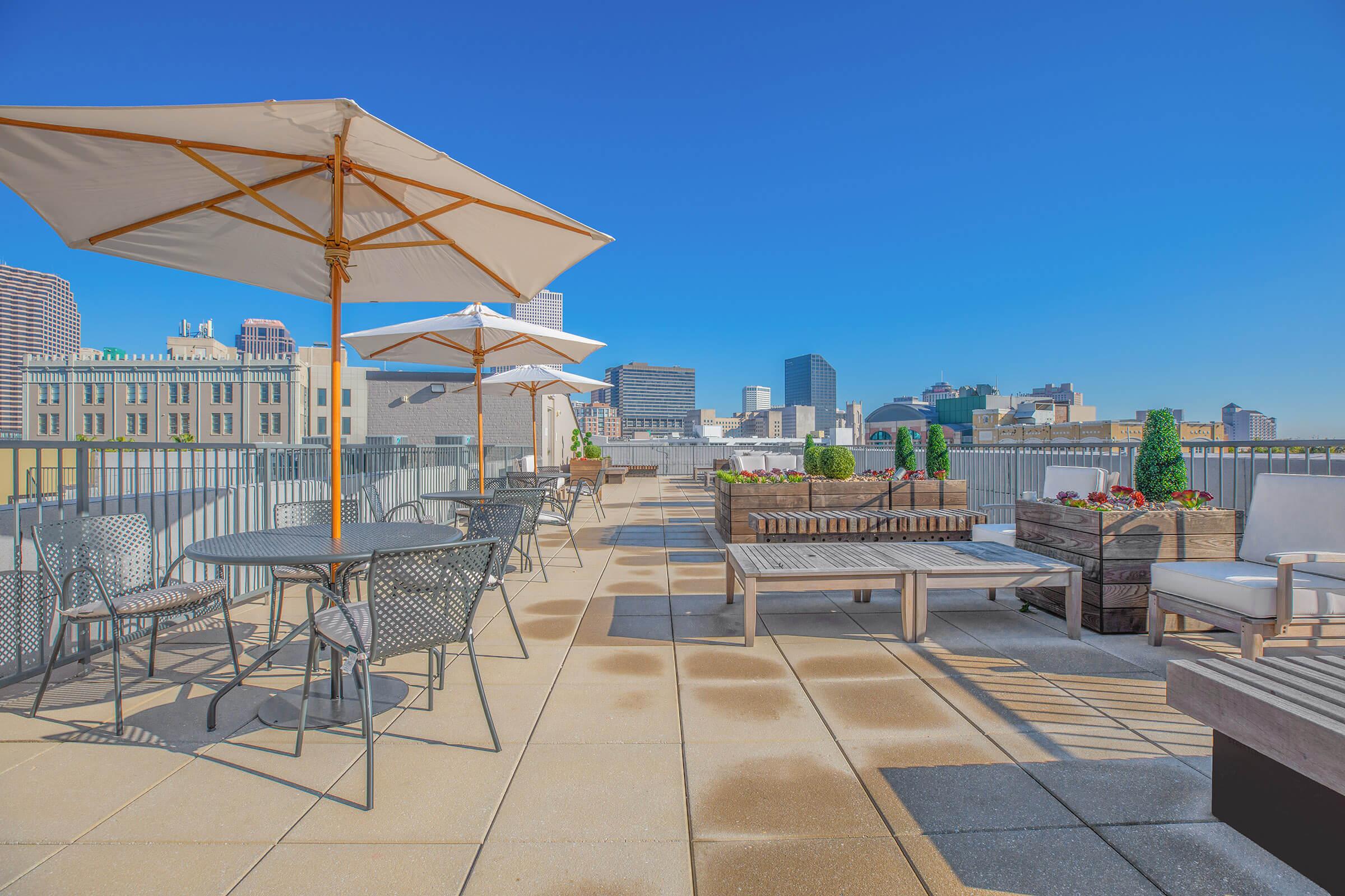 A rooftop terrace featuring modern outdoor furniture, including tables and lounge seating, shaded by large umbrellas. Planter boxes with greenery are present, and a clear blue sky complements the city skyline in the background, creating a relaxing urban retreat.