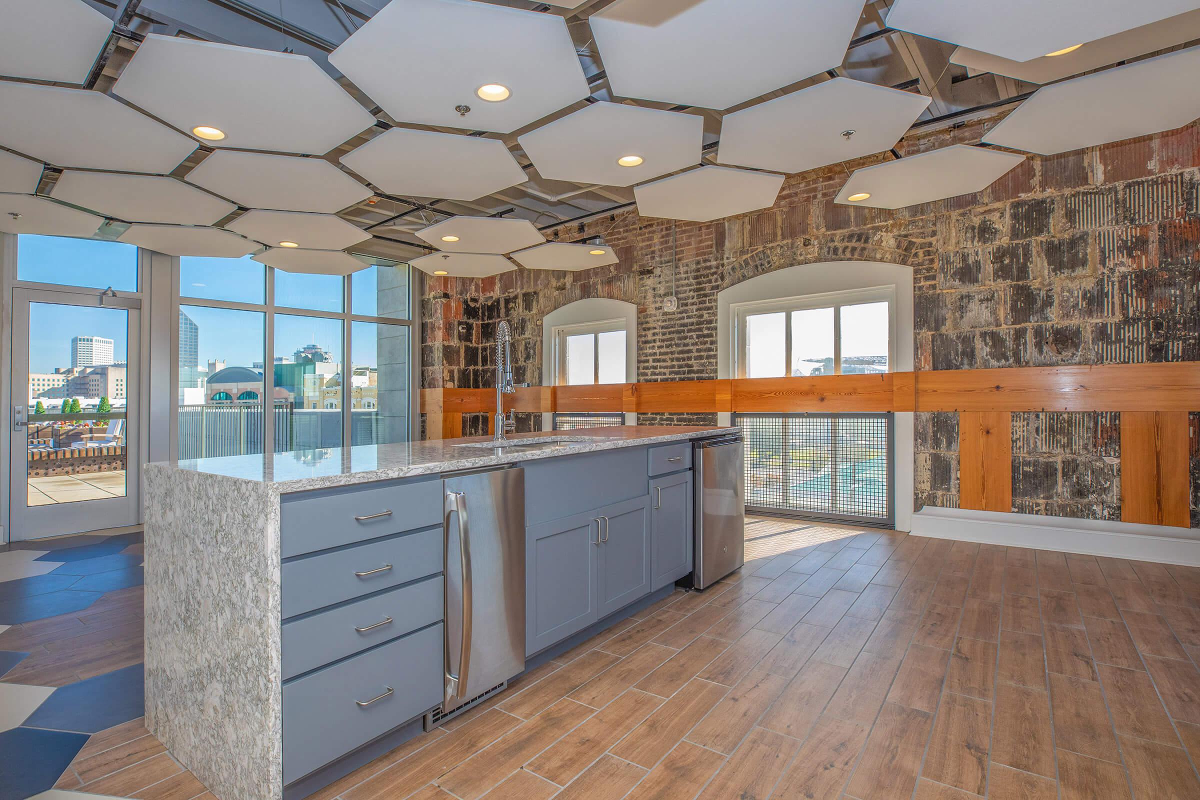 Modern kitchen interior featuring a large island with a gray countertop, stainless steel appliances, and wooden cabinetry. The room has unique hexagonal ceiling tiles, large windows providing natural light, and a stylish mix of brick and wood accents in the background.