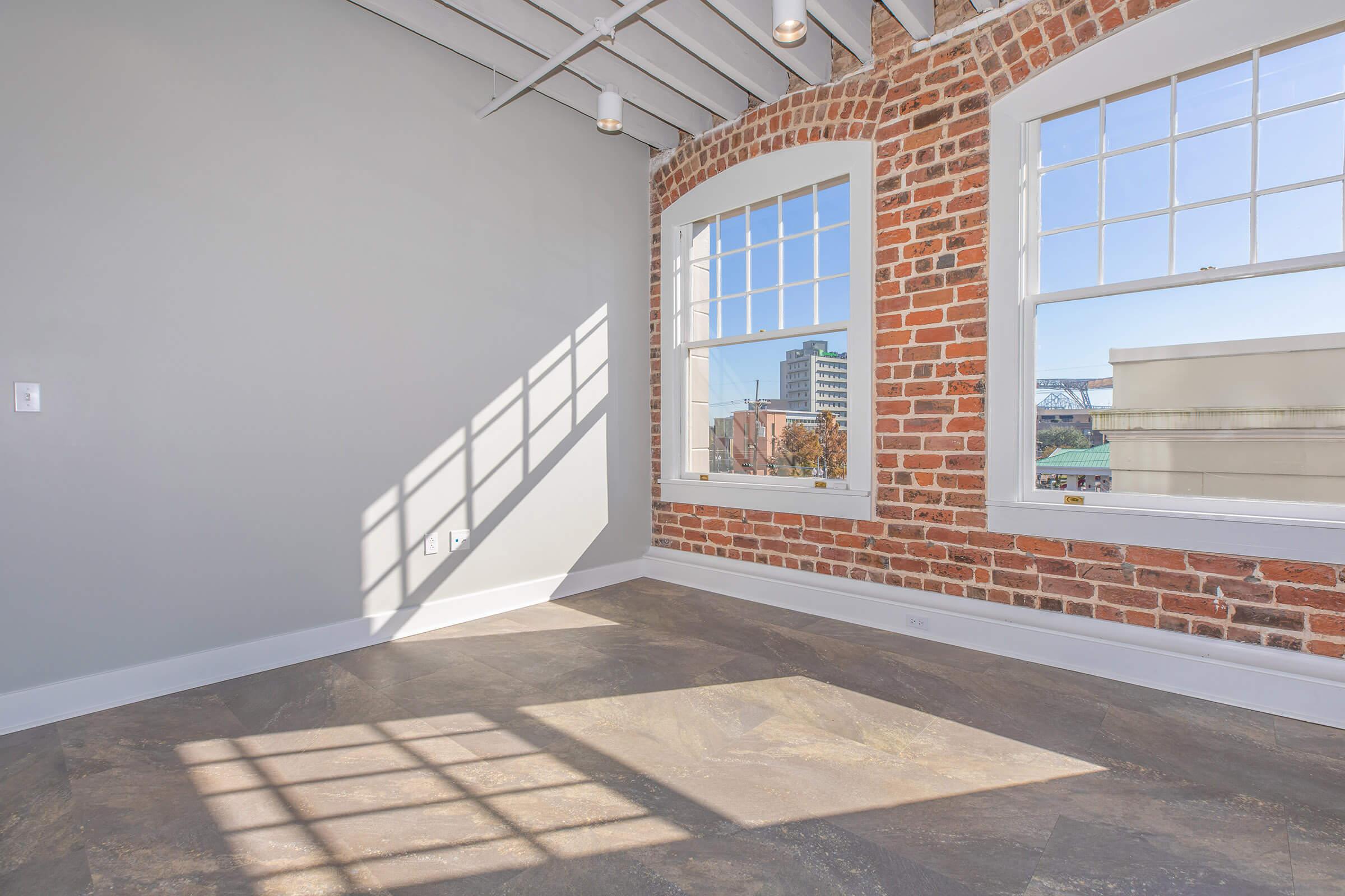 A bright, modern room with exposed brick walls and large windows allowing natural light to enter. The room features a gray wall and a polished floor, with shadows cast from the windows creating an interesting pattern on the floor. The view outside includes urban buildings.