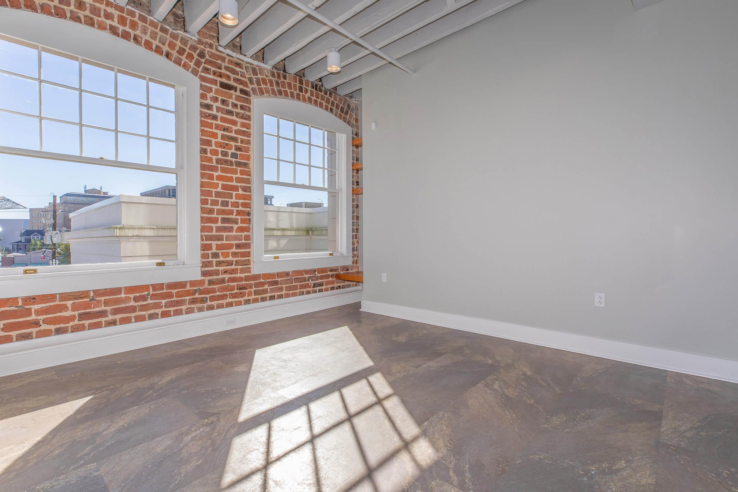 A well-lit, modern room featuring exposed brick walls and large windows. The flooring is made of grey tiles, with sunlight casting geometric shadows on the surface. The ceiling is adorned with beams, creating an airy and spacious atmosphere.