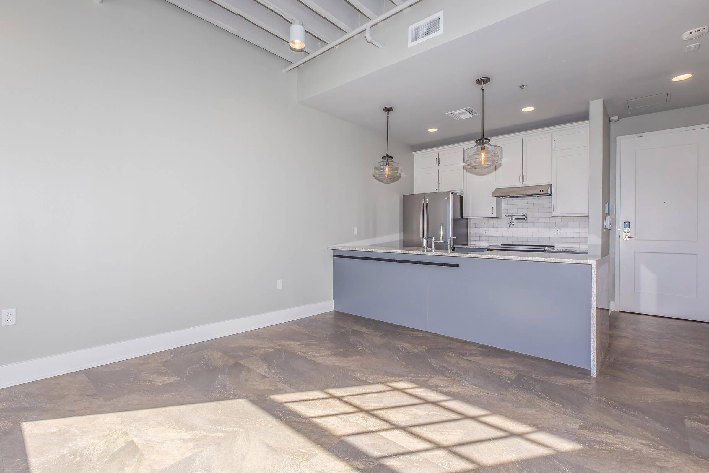 Modern kitchen and living area with light gray walls and a concrete floor. Features an island with a dark blue base and white marble countertop, stainless steel appliances, and pendant lighting. Large windows create natural light, casting shadows on the floor. Sleek design with minimalist aesthetics.