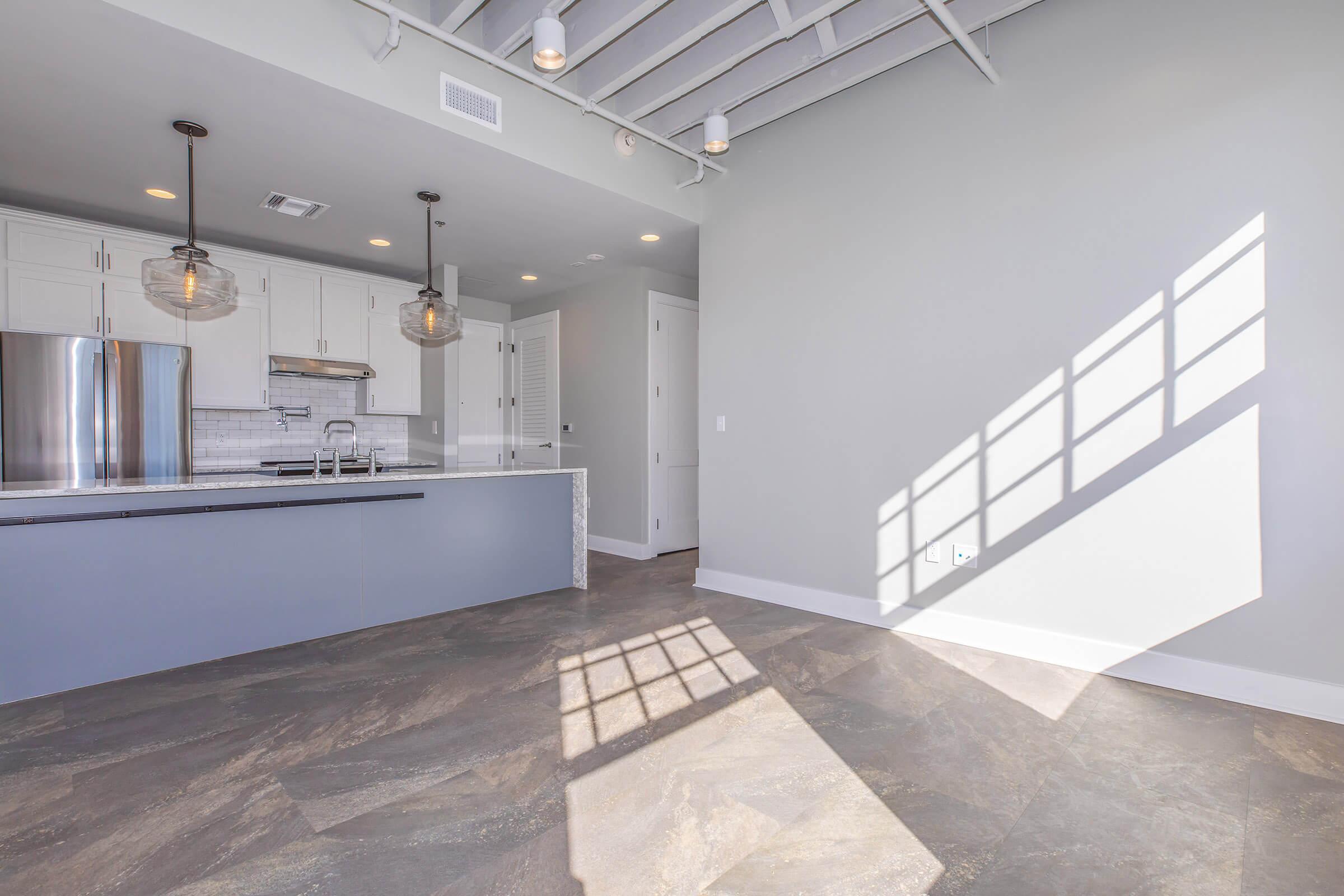 A modern kitchen and living area with white cabinetry, stainless steel appliances, and gray walls. The sunlight casts a geometric shadow from a window onto the floor, creating a warm ambiance. The space features an open layout, highlighting contemporary design elements.