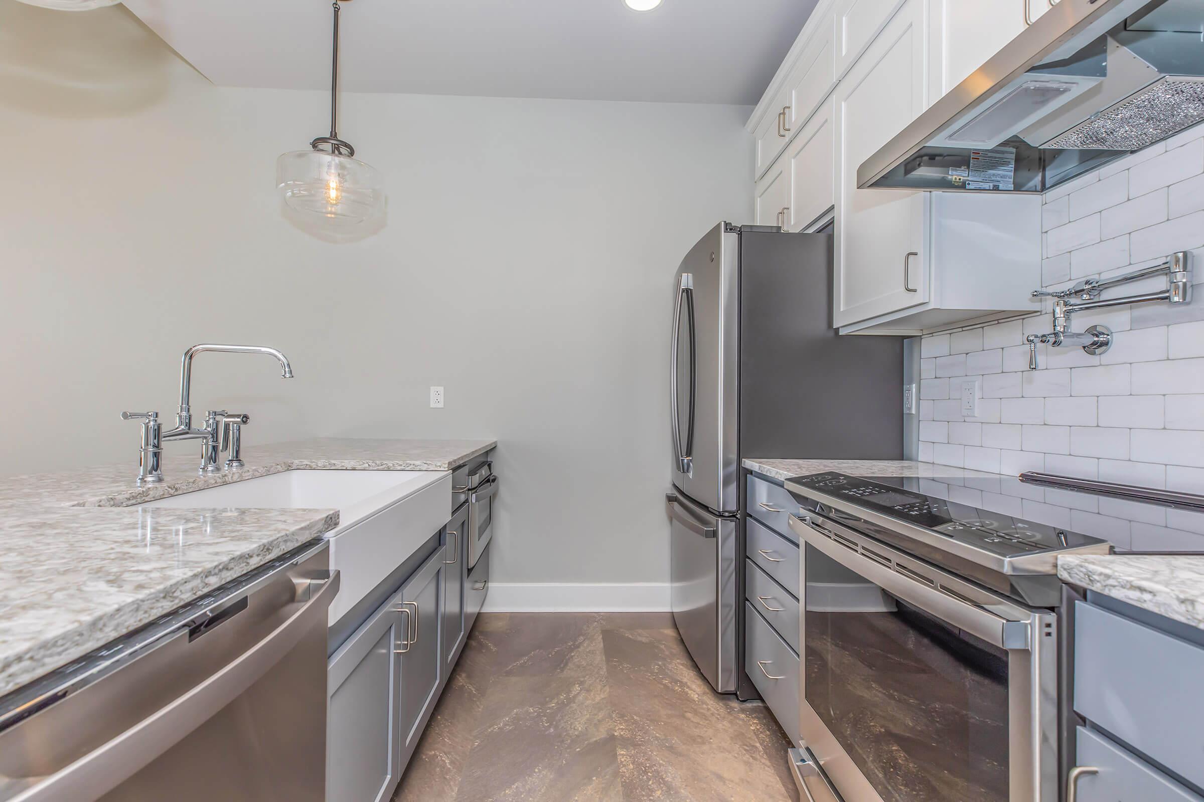 A modern kitchen featuring stainless steel appliances, including a refrigerator, stove, and dishwasher. The countertops are made of gray granite, and there is a white farmhouse sink. The cabinetry is light gray, and a pendant light hangs from the ceiling, adding a stylish touch to the space. The backsplash is white subway tile.