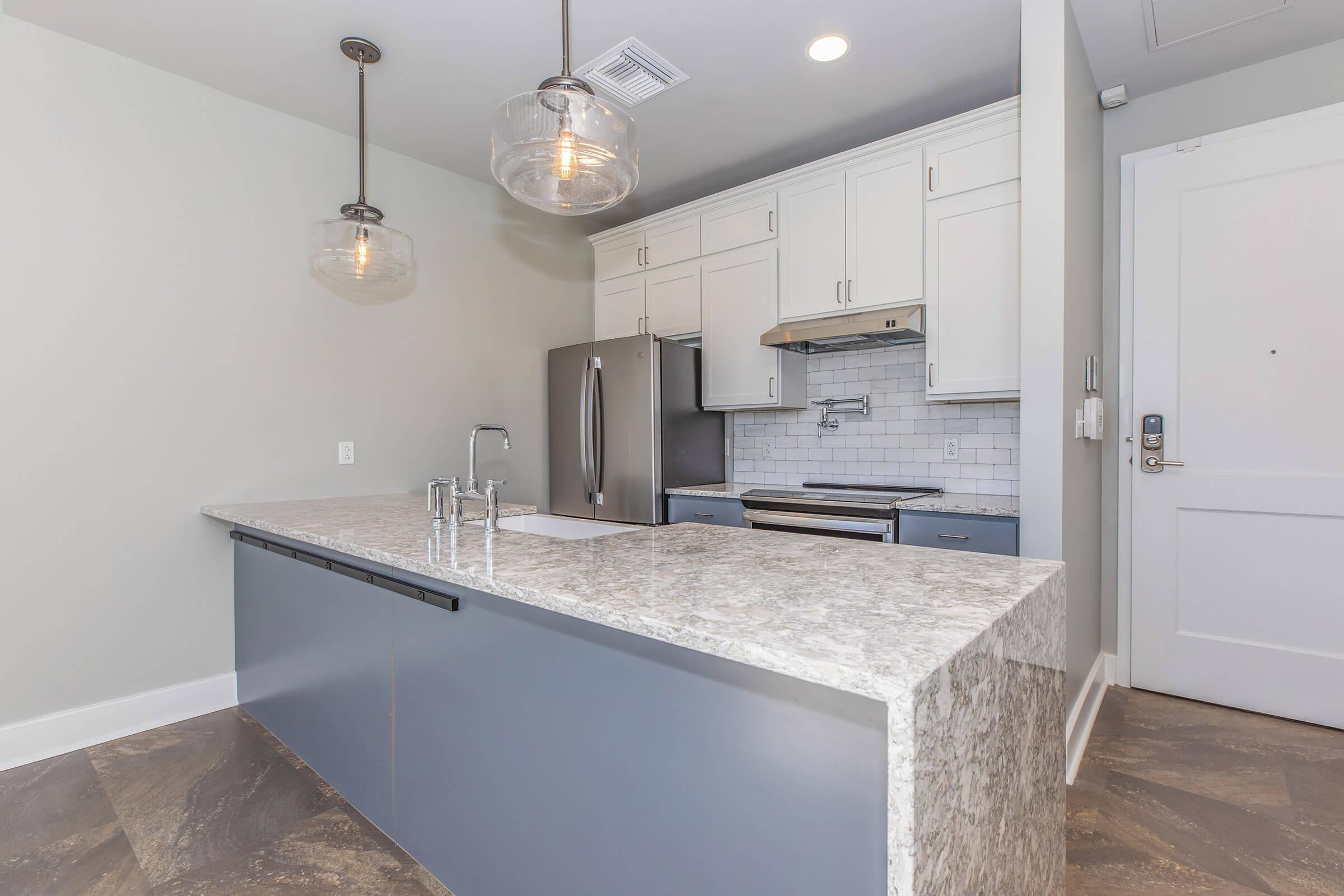 A modern kitchen featuring light gray walls, a large island with a speckled granite countertop, stainless steel appliances, and pendant lighting. The cabinets are white with a sleek design, and there is a silver faucet over a stainless steel sink. The flooring is dark, adding contrast to the space.