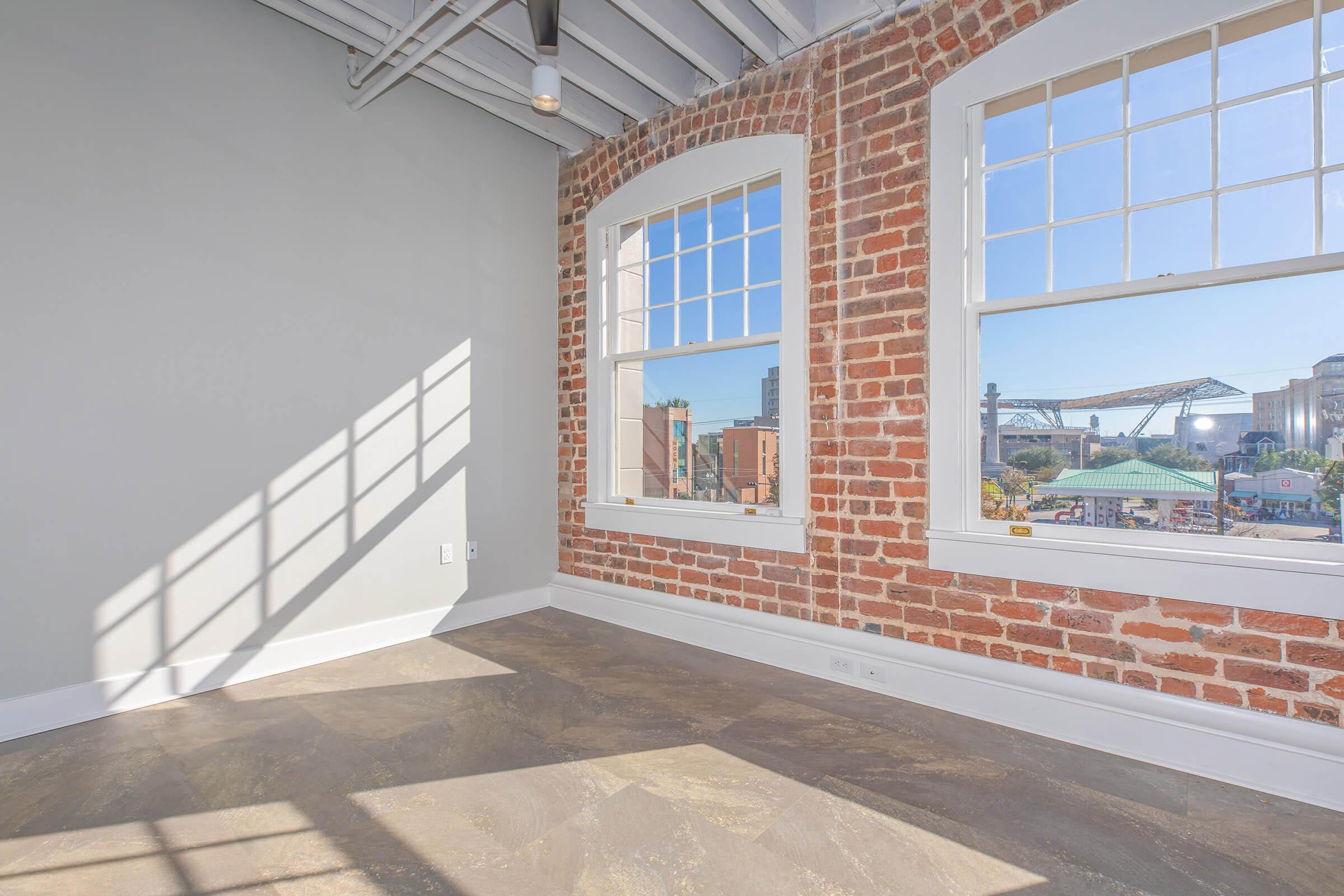 A bright, empty room with exposed brick walls and large windows allowing natural light to flood in. The floor is a light-colored material. Outside the windows, there are glimpses of a scenic urban landscape, including buildings and a structure in the distance.