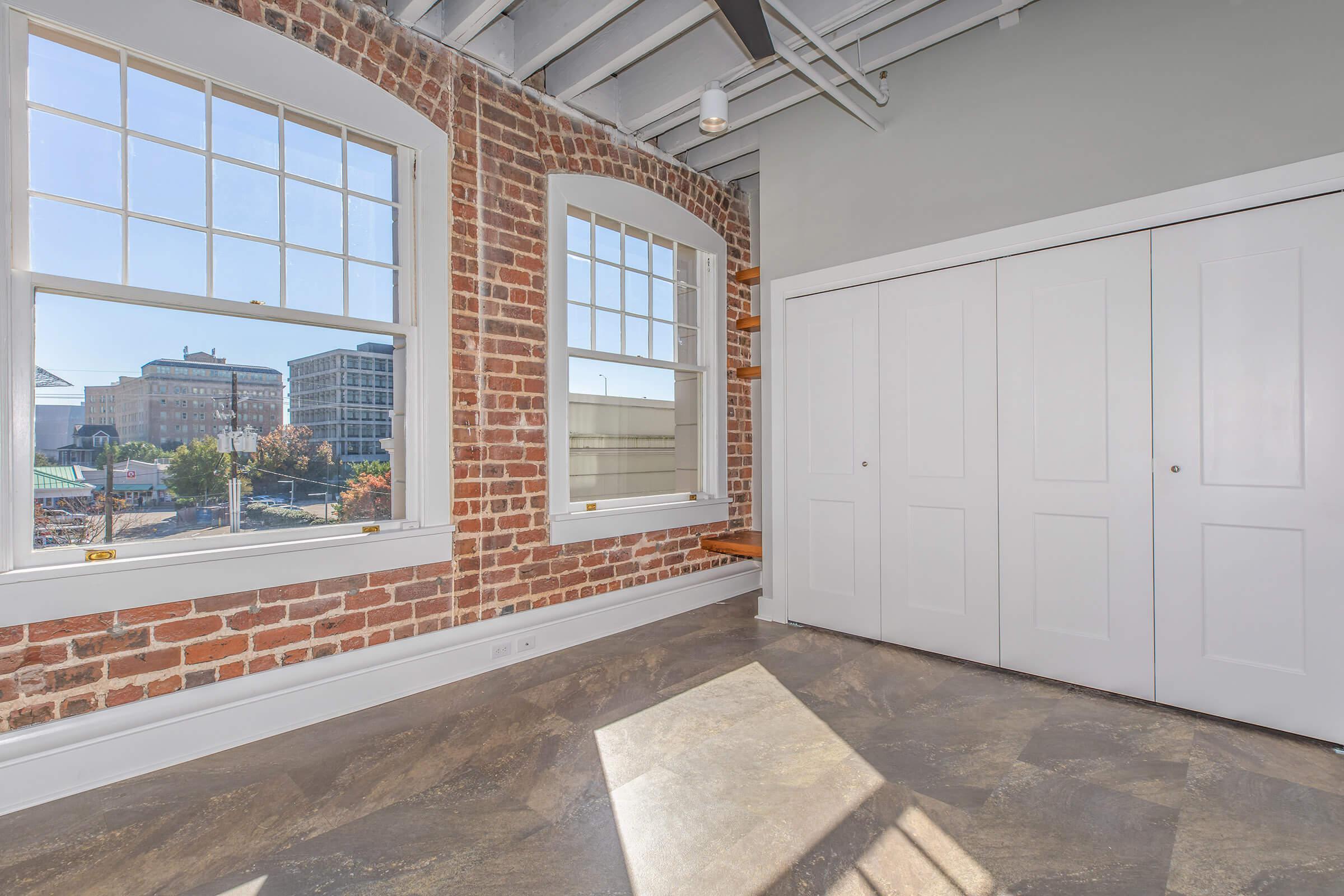 Interior of a sunny room featuring exposed brick walls, large windows allowing natural light, and a light-colored floor. There is a double closet with white sliding doors on one side, and a view of buildings outside the windows. The overall style is modern with industrial elements.