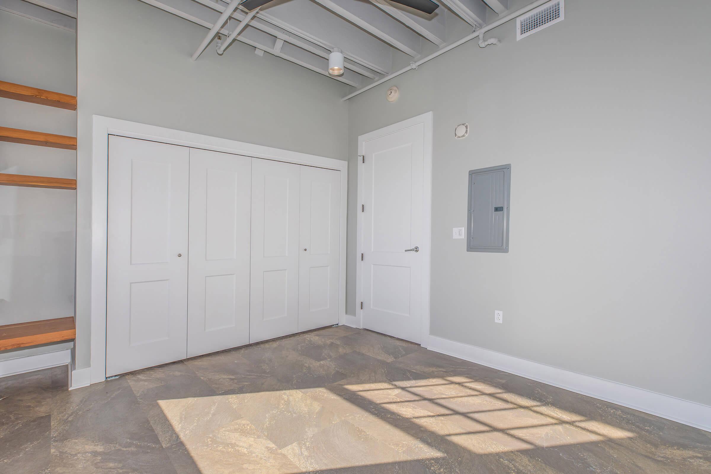 A modern, empty room featuring light gray walls, a beige tiled floor, and a large closet with double doors. There are white baseboards, a wall-mounted electrical panel, and a wooden shelf against one wall. Bright sunlight filters in, creating shadows on the floor.