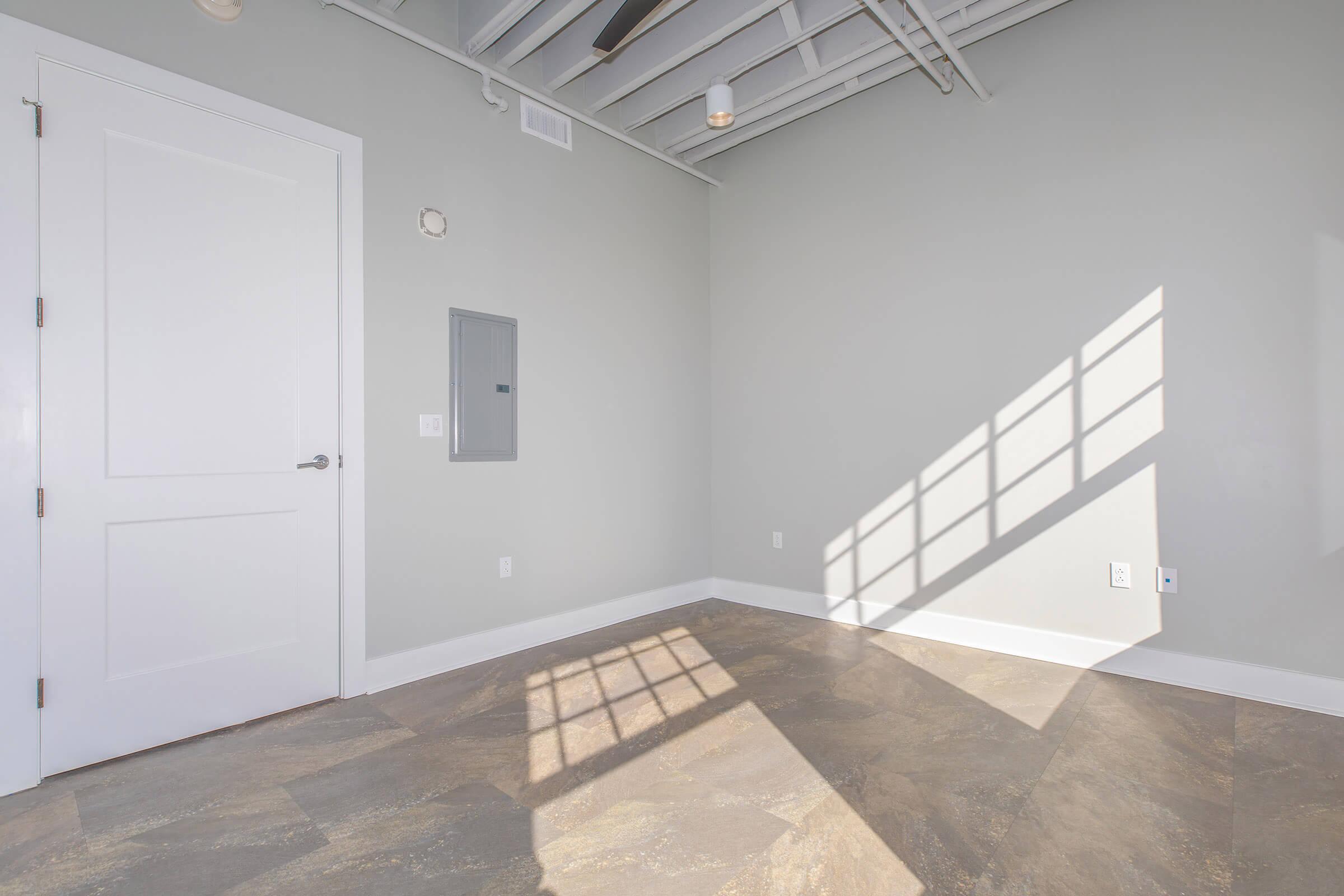 An empty room with gray walls and a textured floor. A white door is visible on the left, while a gray electrical panel is mounted on the wall. Bright sunlight casts a shadow across the floor from a window, creating a pattern on the surface. The ceiling features exposed beams.