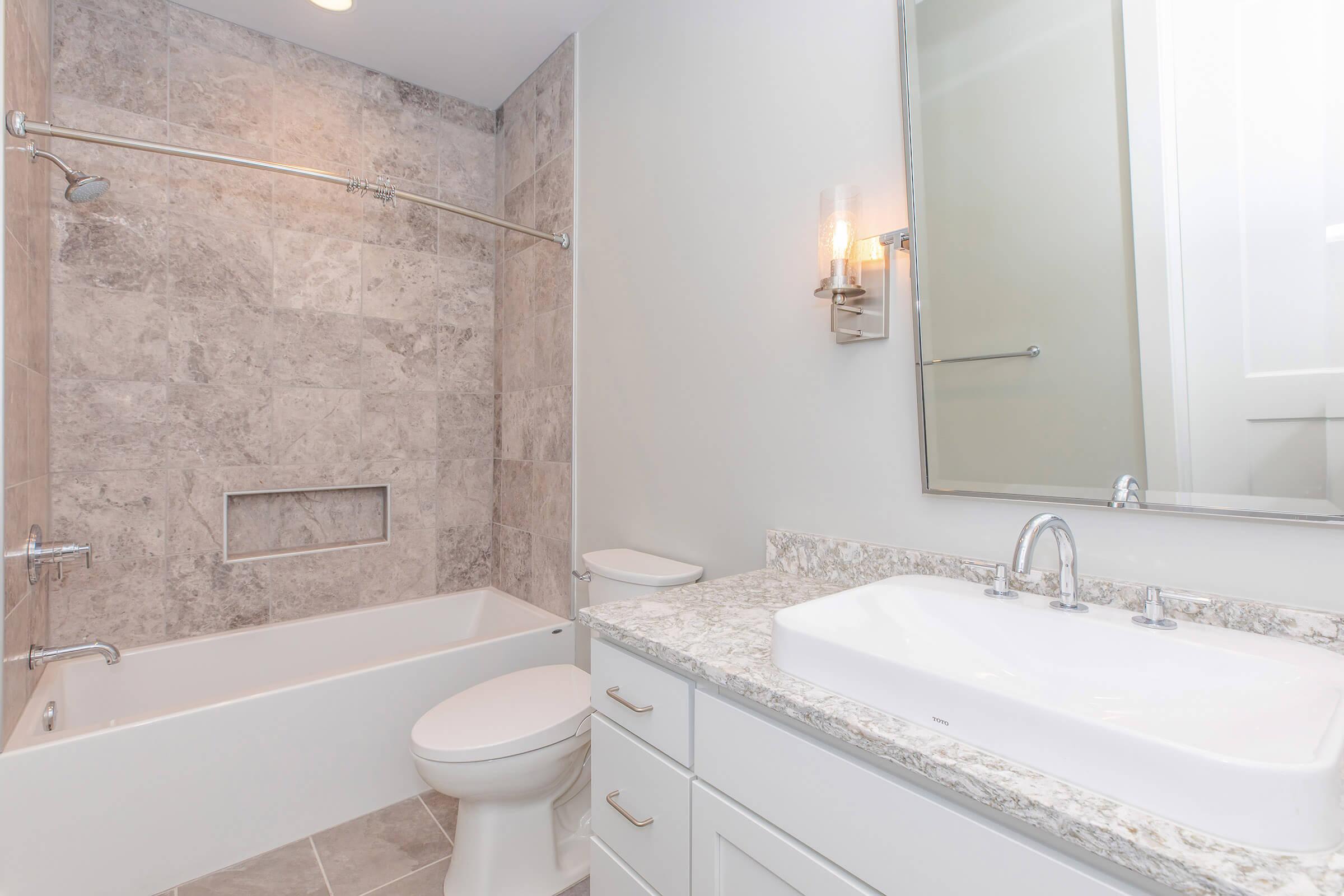 Modern bathroom featuring a light gray tiled wall, a white bathtub with a glass shower rod, and a sleek double sink vanity with granite countertop. There's a wall-mounted mirror above the sink and a stylish light fixture nearby, creating a bright and clean atmosphere.