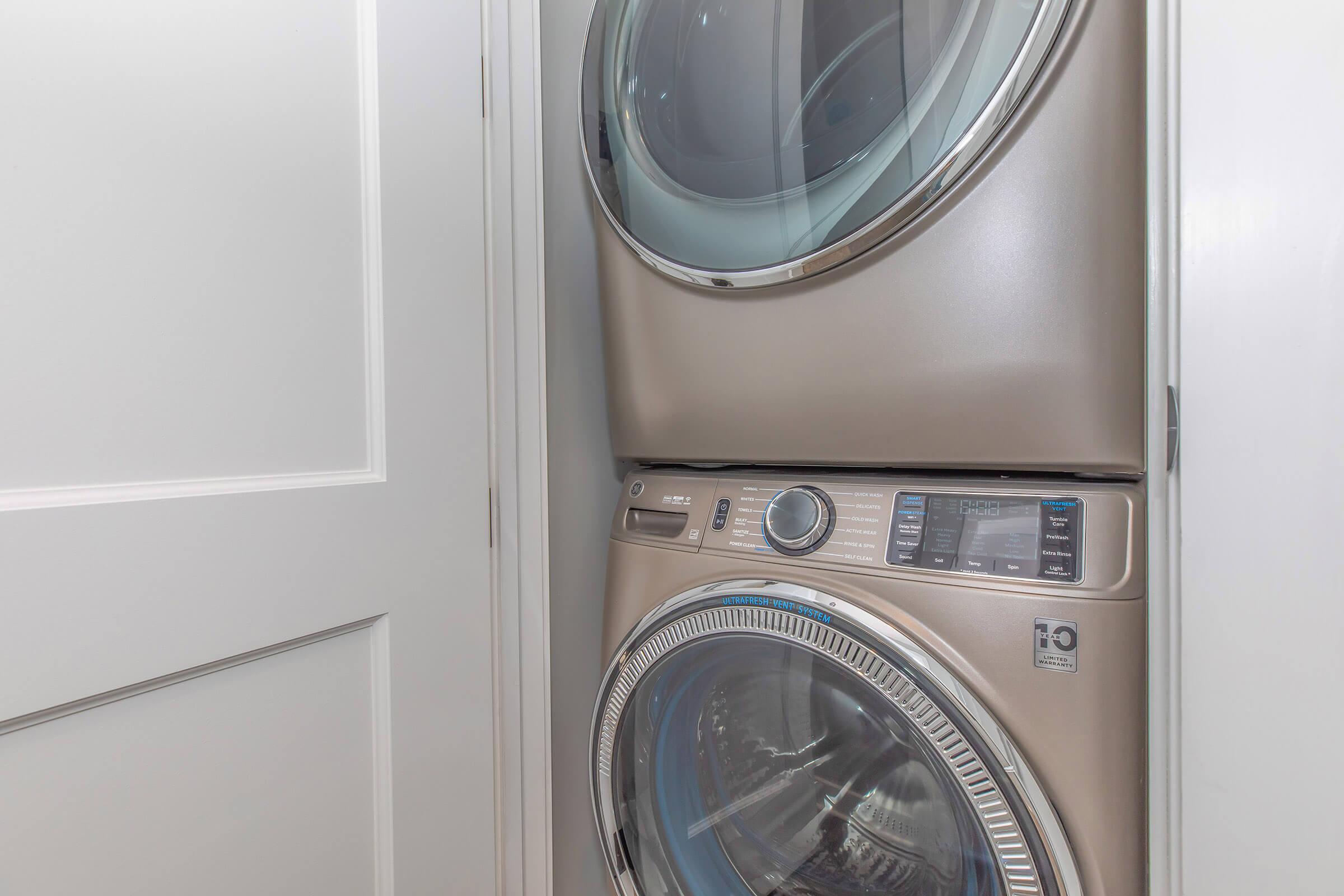 A stacked washer and dryer in a laundry room, featuring a modern design with a sleek metallic finish and large circular doors. They are positioned side by side against a white wall, with a door partially visible in the background. The control panel displays various settings and options for laundry use.