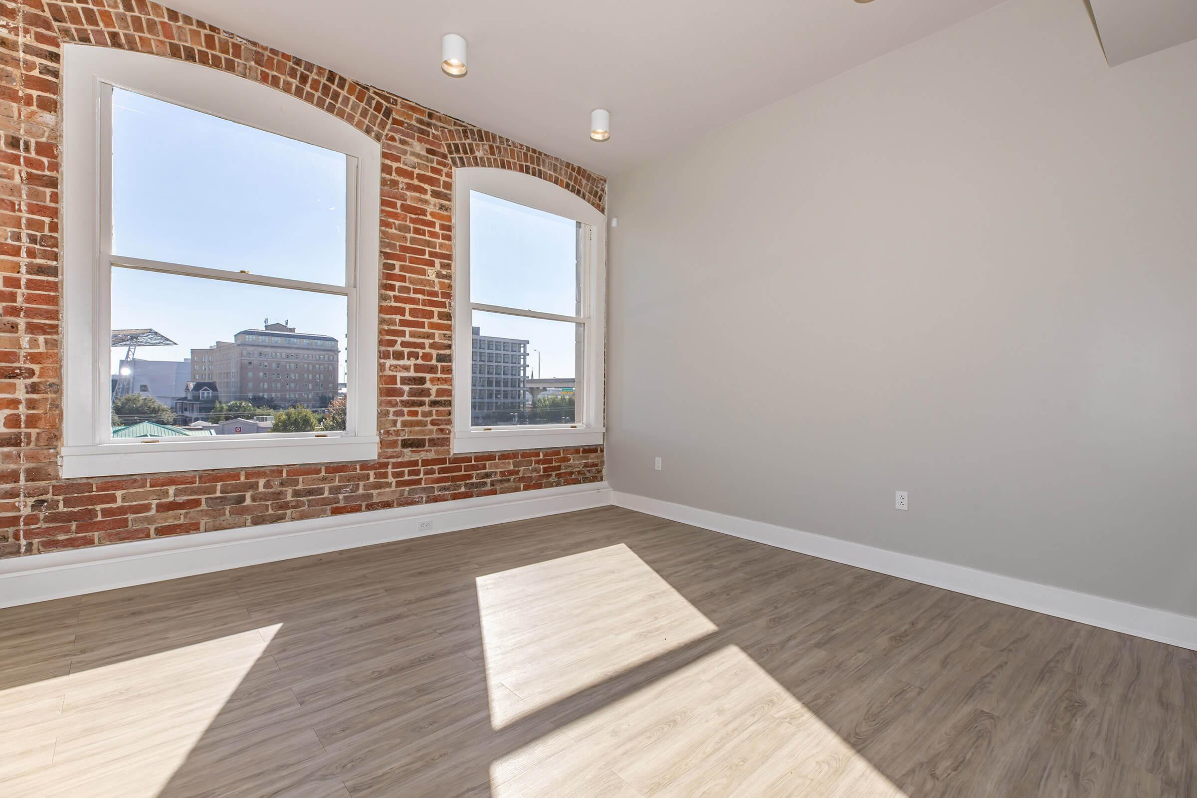 Bright, spacious room with large windows showcasing a view of buildings outside. Exposed brick walls and light-colored flooring create a modern yet cozy atmosphere. Sunlight casts shadows on the floor, highlighting the open layout and inviting natural light into the space.