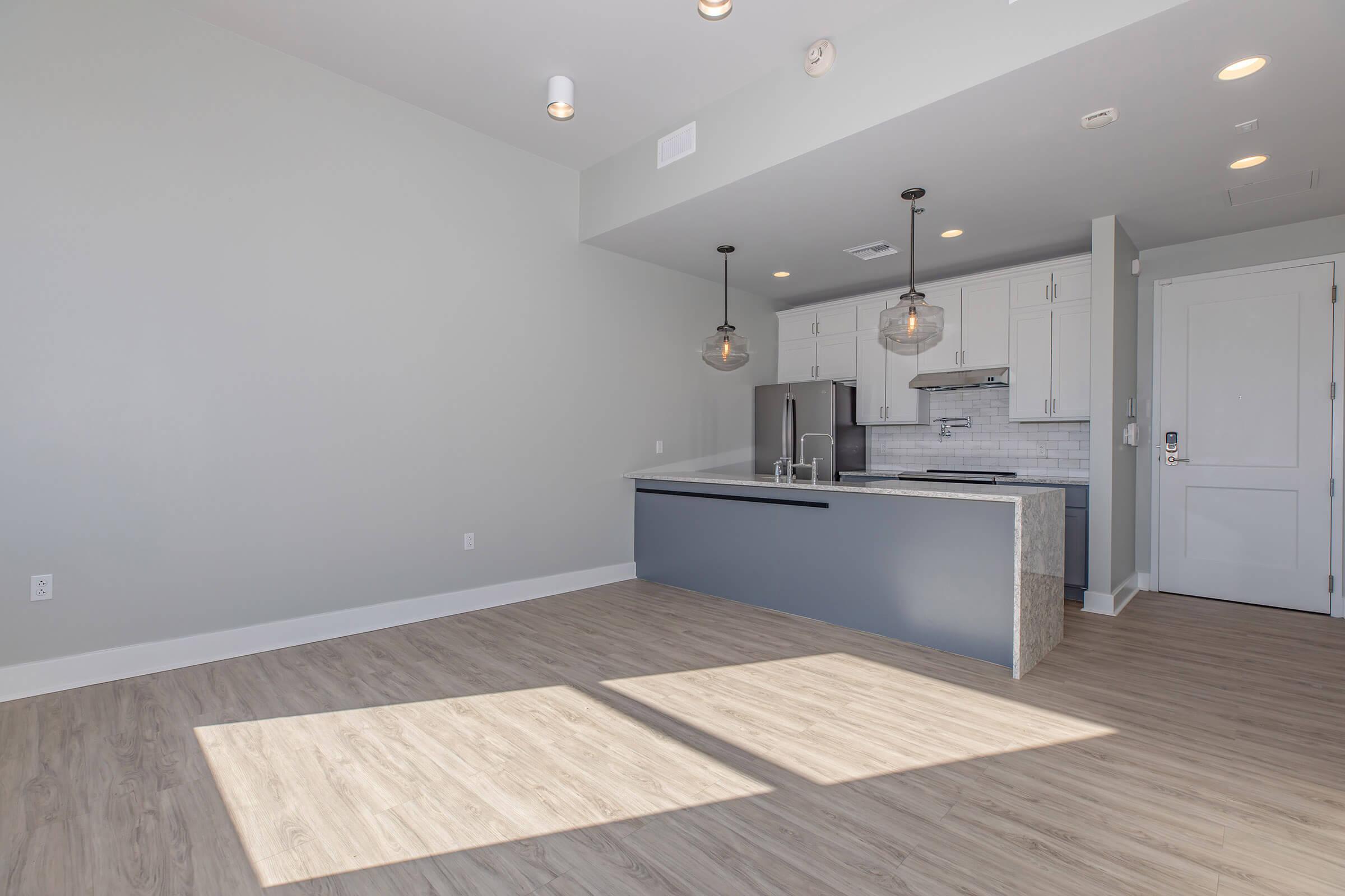 An empty modern kitchen and living space with wooden floors, featuring pendant lighting above a kitchen island, stainless steel appliances, and a light gray color scheme. Large windows allow natural light to flood the room.