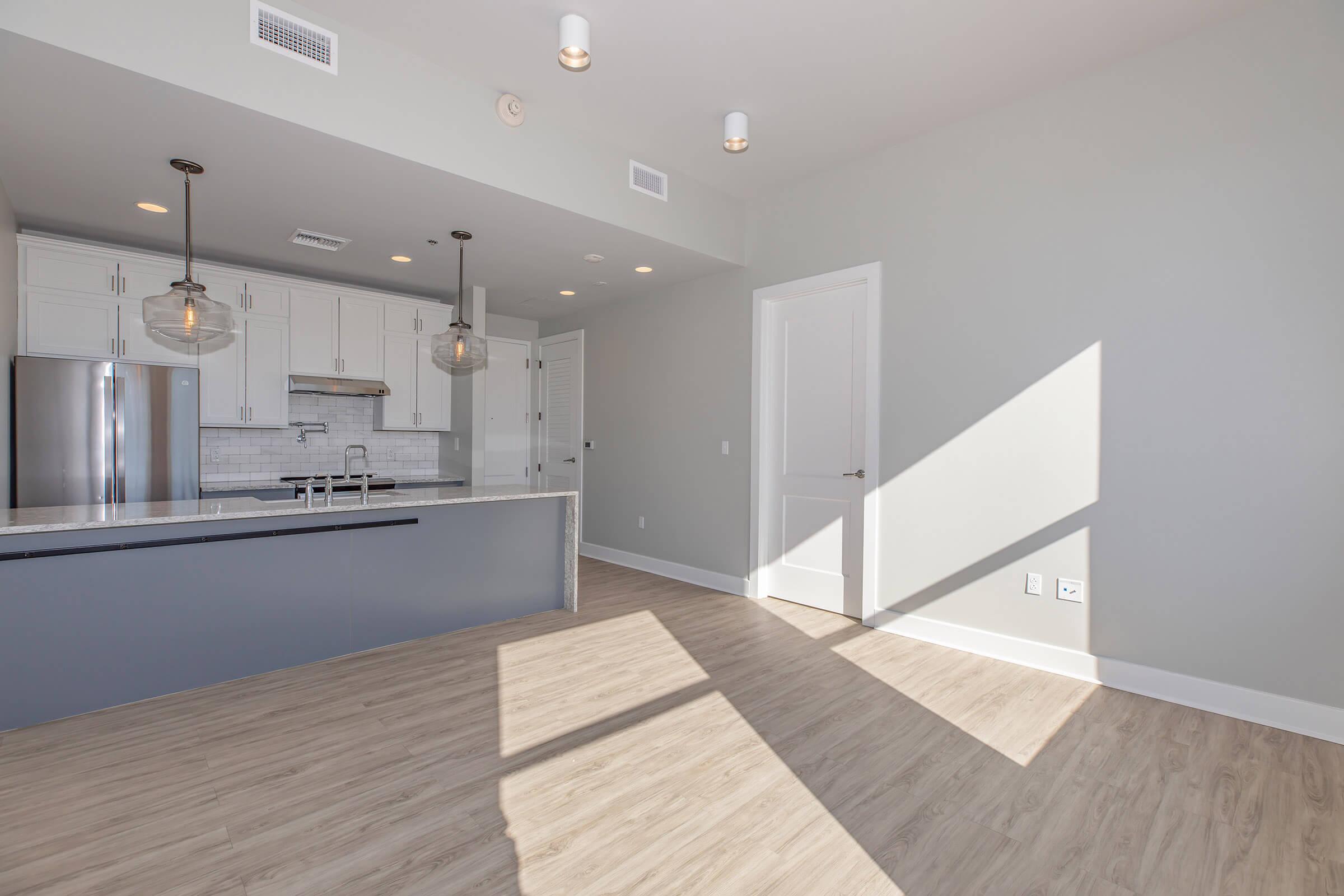 A modern kitchen and living area featuring light gray walls, sleek white cabinetry, stainless steel appliances, and a large kitchen island with a marble countertop. Natural light streams in through a window, casting shadows on the light wooden floor. Bright and airy space with pendant lighting and a door leading to another room.