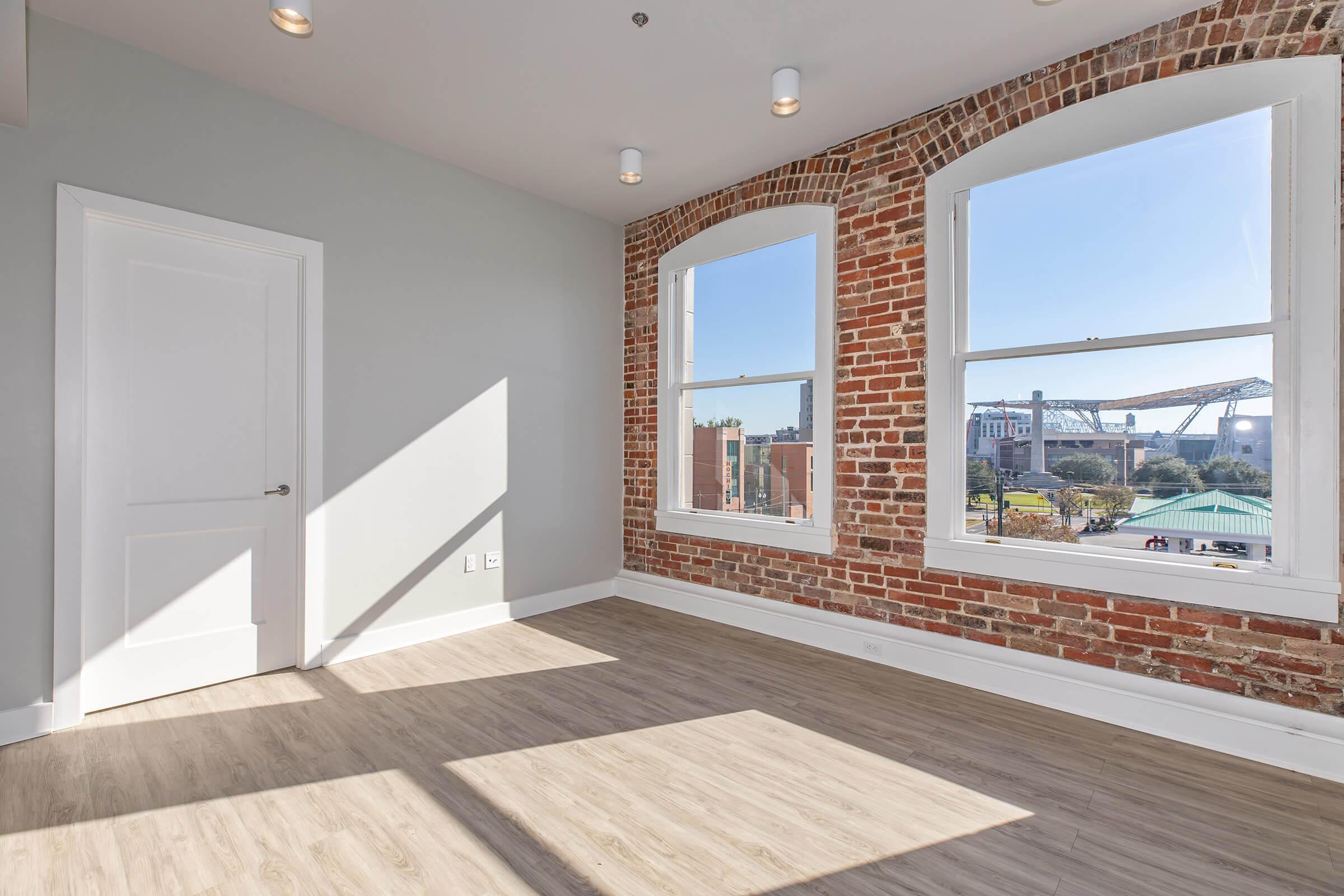 A bright, modern room with large windows showcasing a view of a bridge. The walls feature exposed brick and a light gray paint, while the floor is composed of wooden planks. Natural light floods the space, creating a warm and inviting atmosphere. The door is white and sleek, complementing the overall aesthetic.