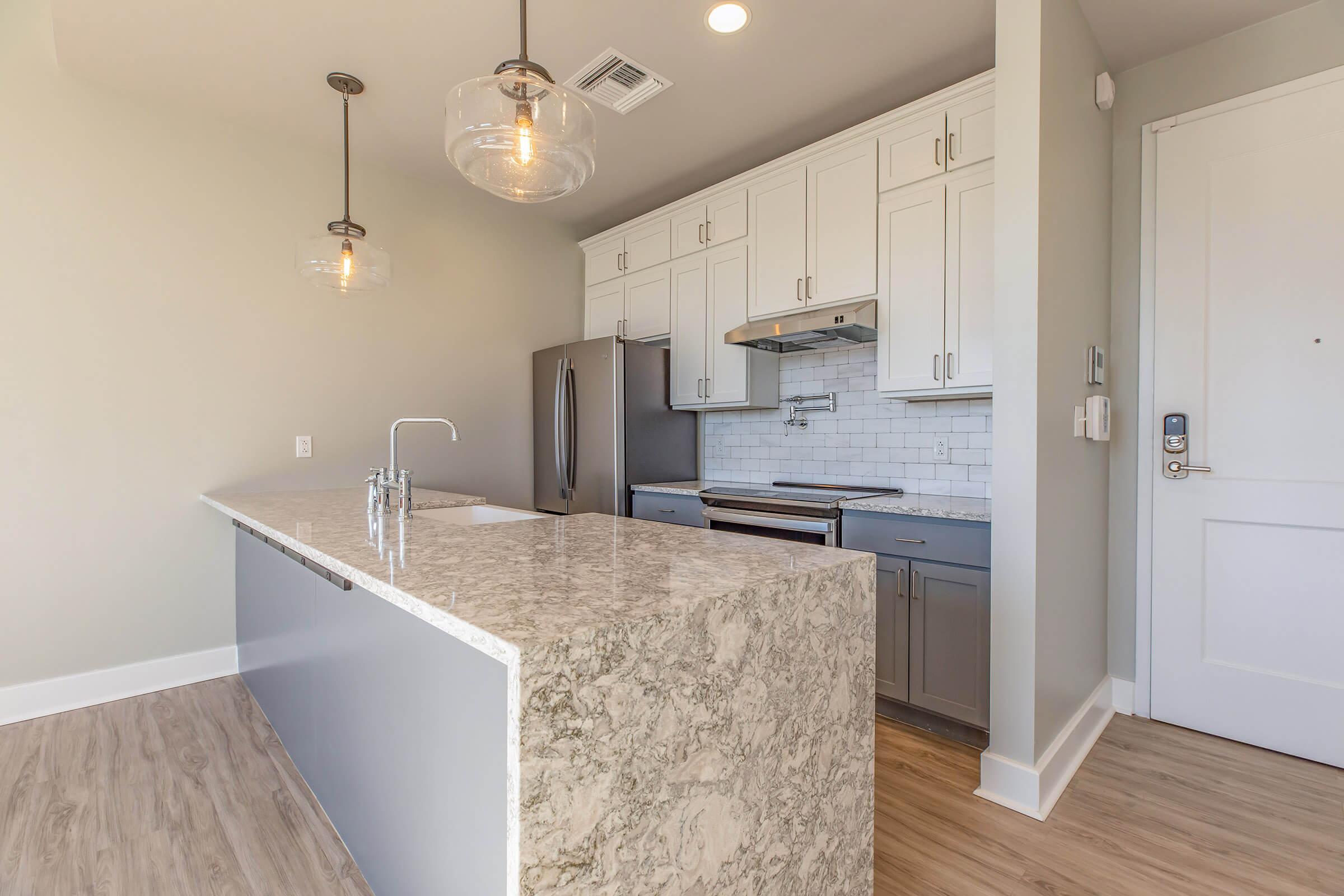 A modern kitchen featuring a large island with a gray and white granite countertop, stainless steel appliances, and white cabinetry. The space has pendant lighting and a neutral-colored wall, giving it a bright and airy feel, with a door leading to the exterior.