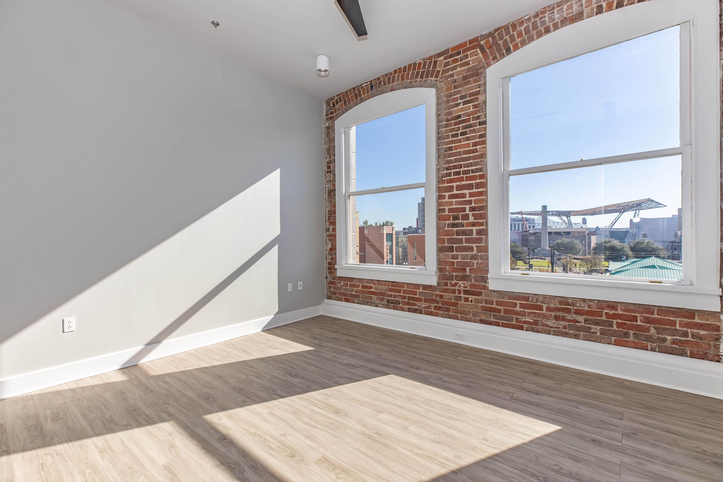 A brightly lit room with large windows showcasing a view outside. The walls feature exposed brick, while the floor is made of light-colored wood. Sunlight casts a shadow across the room, creating a warm and inviting atmosphere. The space appears modern and empty, ideal for potential furnishing.