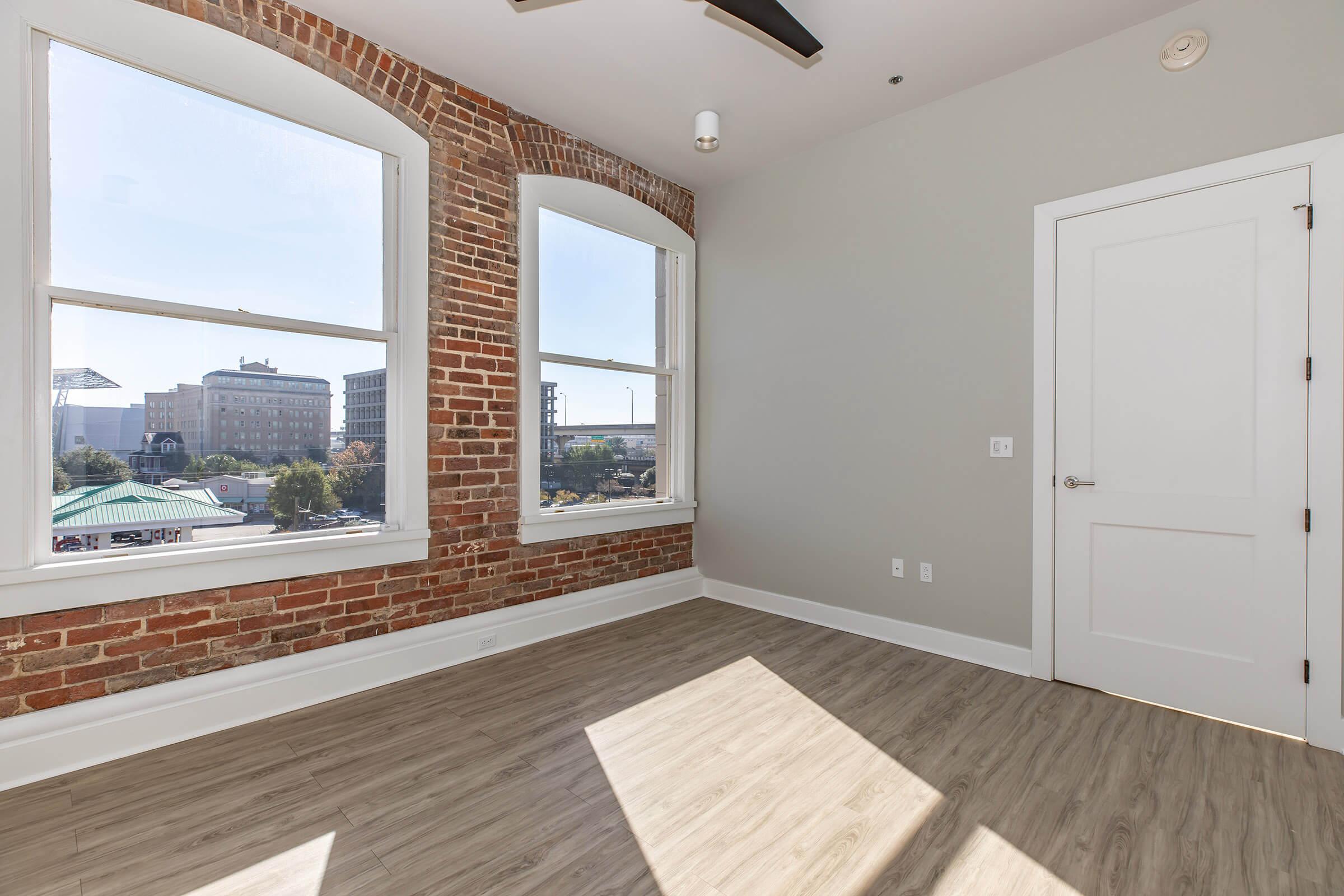 A bright, modern room featuring exposed brick walls and large windows allowing natural light. The floor is made of light-colored wood, and it has a simple white door against a light gray wall. The view outside shows a cityscape, enhancing the urban feel of the space.