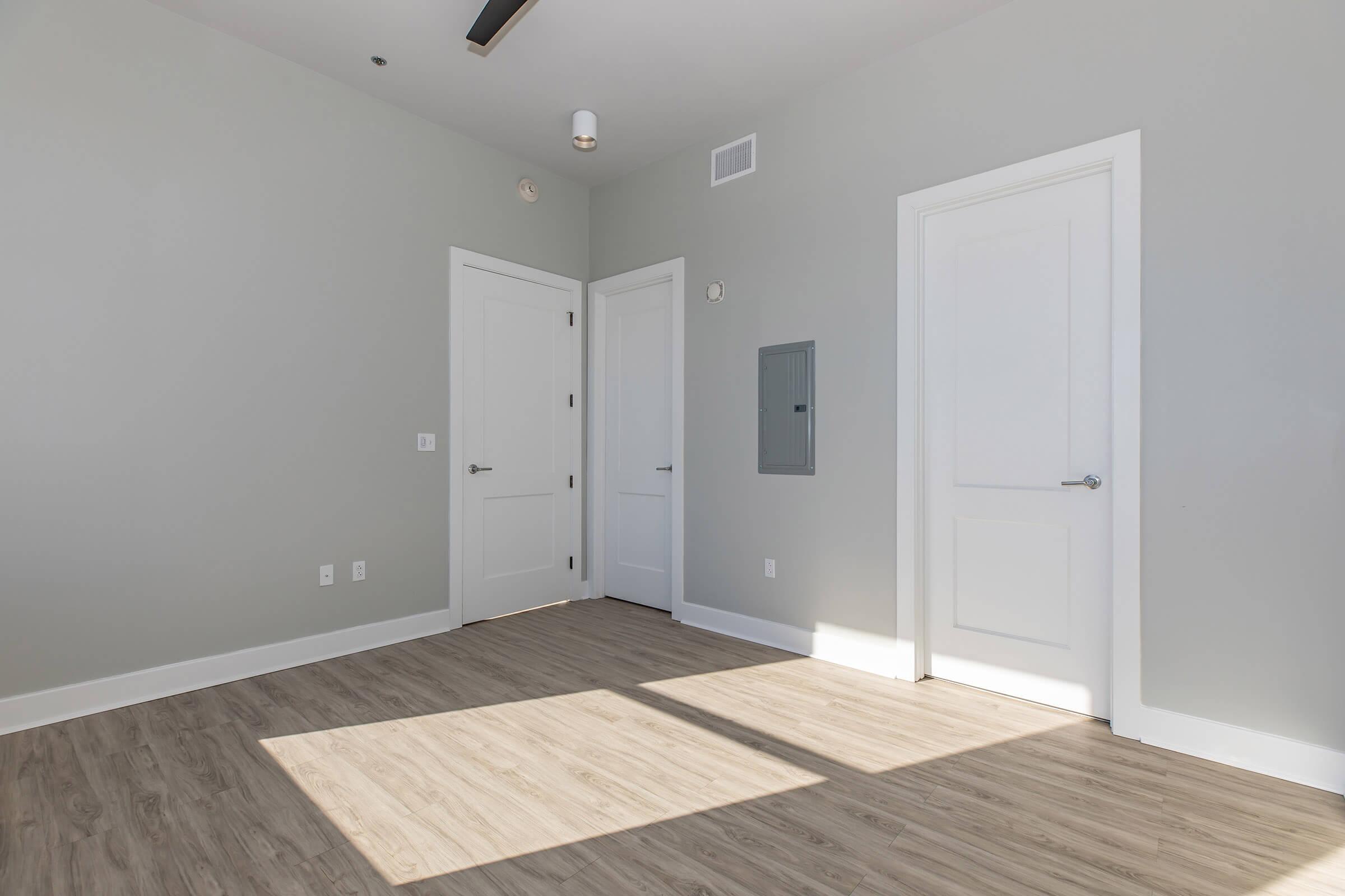 A brightly lit empty room with light gray walls and a light wood floor. Two closed white doors are visible, one leading to the hallway and another to a closet. A small wall panel is present next to the door on the left, and natural light casts a shadow on the floor.