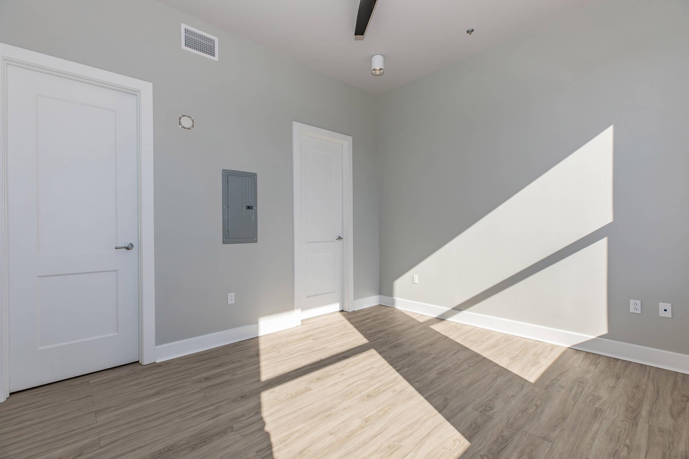 A room with light gray walls and a light wood floor. It features two white doors, one leading to a closet and the other to another room. A wall-mounted electrical panel is visible. Sunlight casts long shadows across the floor, creating a bright and open atmosphere.