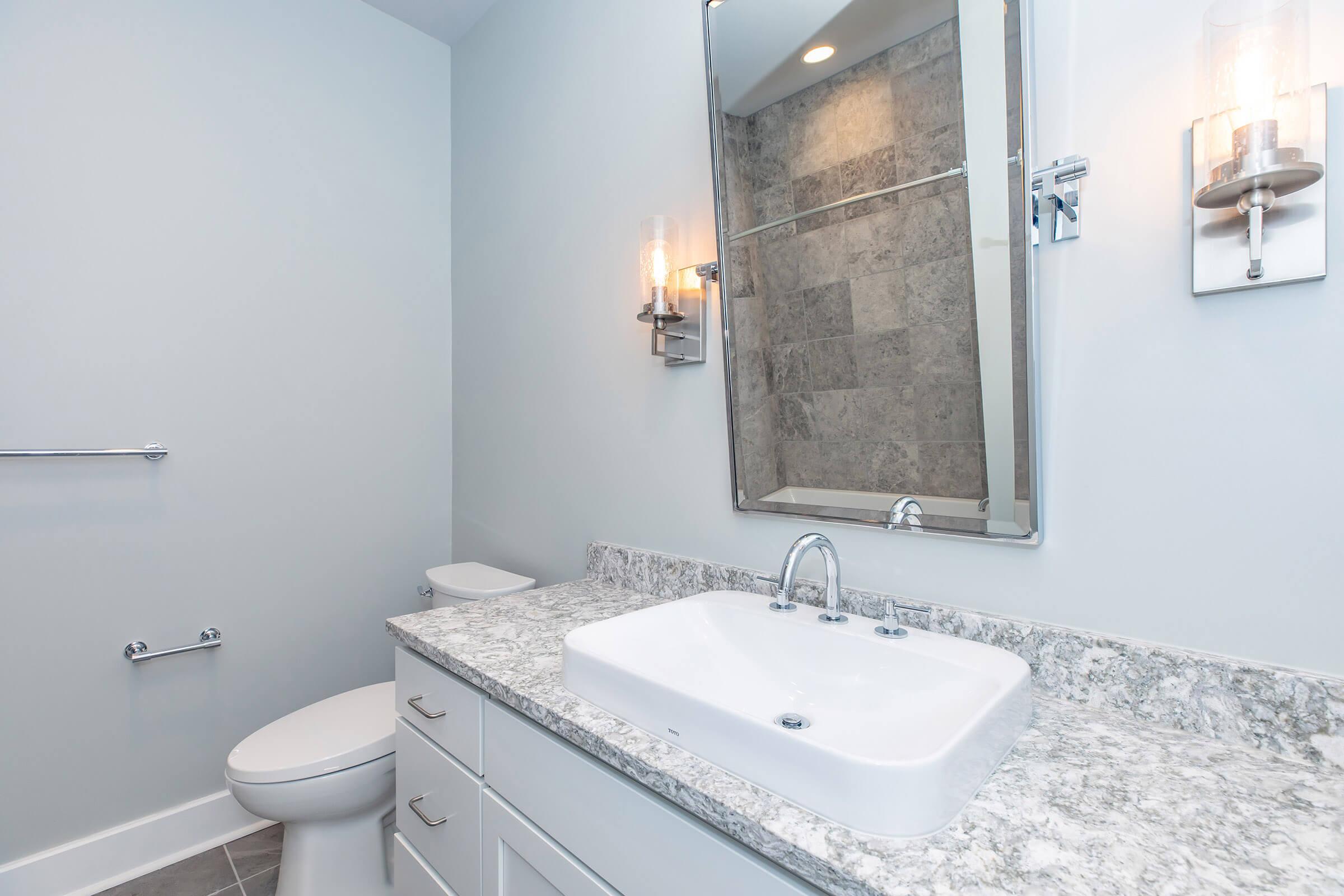 Modern bathroom featuring a white sink on a granite countertop, a wall-mounted mirror with warm lighting, a toilet, and light gray walls. The design is sleek and minimalist, with a tiled shower area visible in the background.