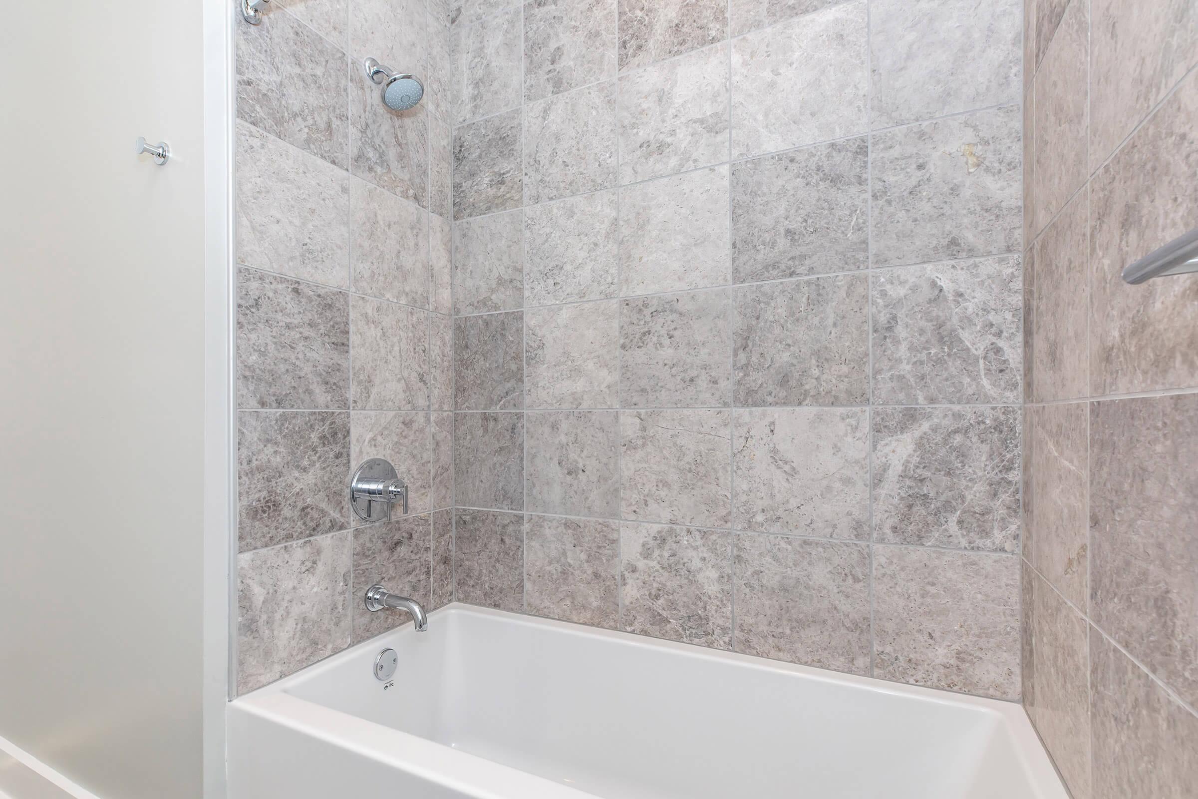 A clean, modern bathroom featuring a white bathtub with a chrome faucet and showerhead. The walls are tiled with large, light gray stone tiles, and the overall design is simple and contemporary, creating a soothing and spacious atmosphere.