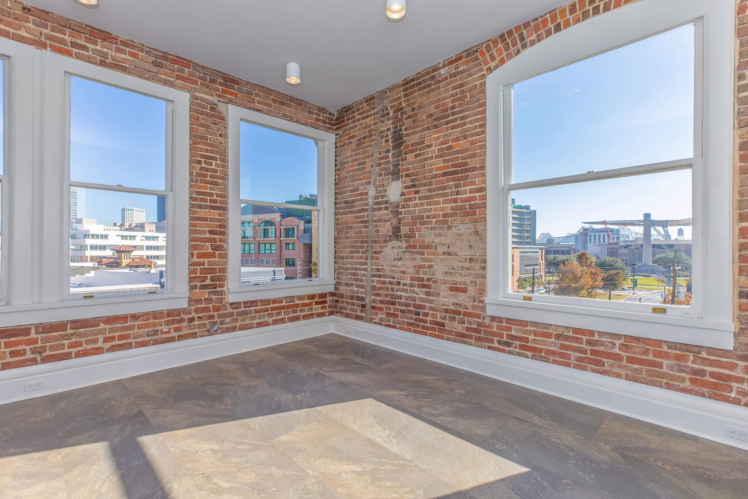 A brightly lit corner room featuring exposed brick walls and large windows that offer a view of the outside urban landscape. The flooring is a mix of tiles, creating a modern yet rustic atmosphere. Natural light fills the space, enhancing the warm tones of the brick.