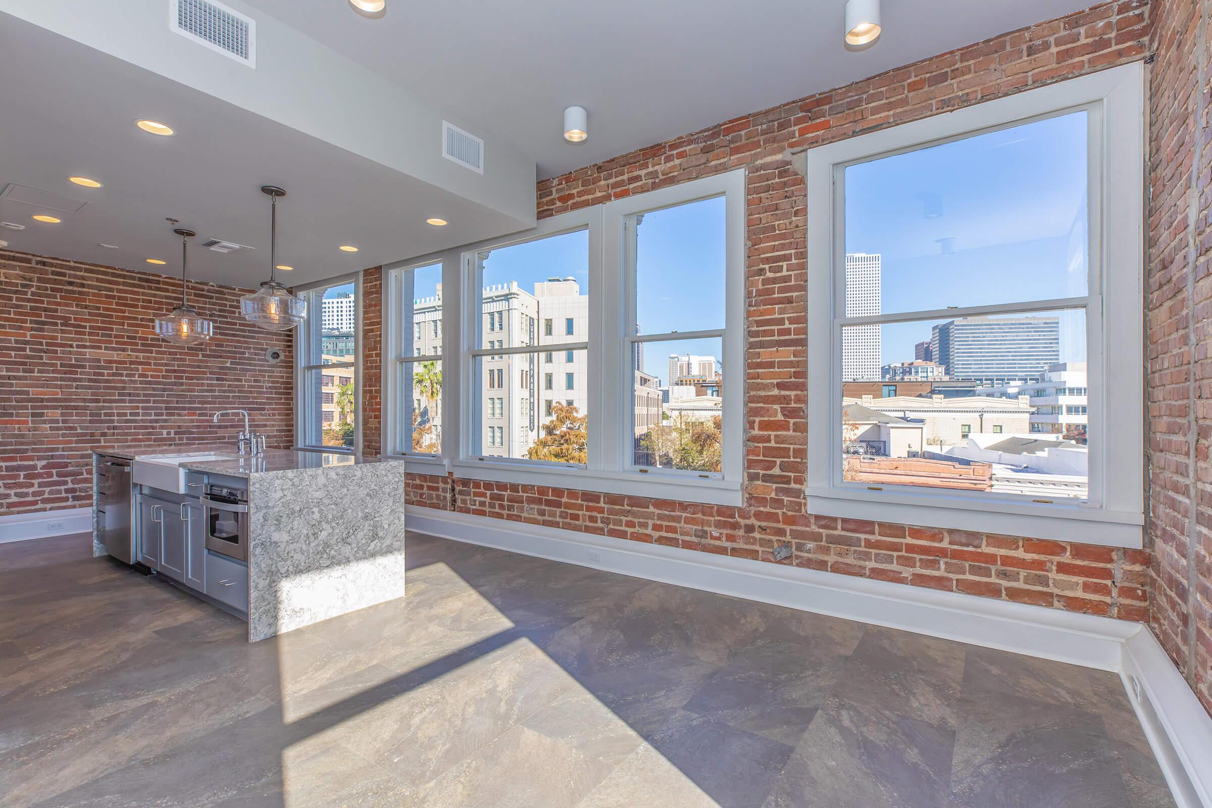 Modern kitchen and living area with exposed brick walls, featuring large windows that provide a view of the city skyline. The kitchen includes a gray marble island and stainless steel appliances, while natural light floods the space, creating an inviting atmosphere.