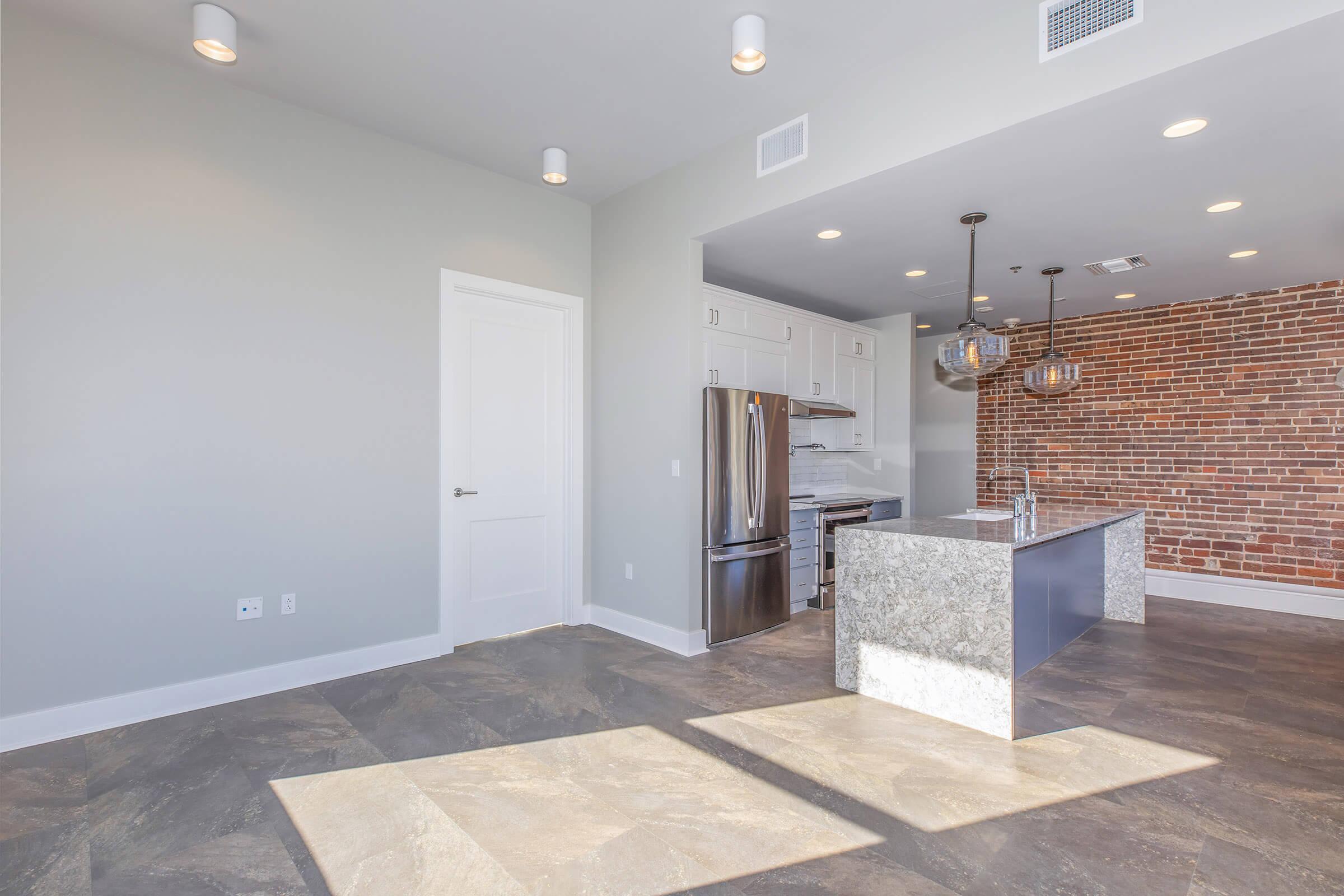 A modern kitchen and living area featuring a sleek island with a granite countertop, stainless steel appliances, and exposed brick accent wall. The space has large windows allowing natural light, contemporary lighting fixtures, and light-colored walls, creating a stylish and inviting atmosphere.