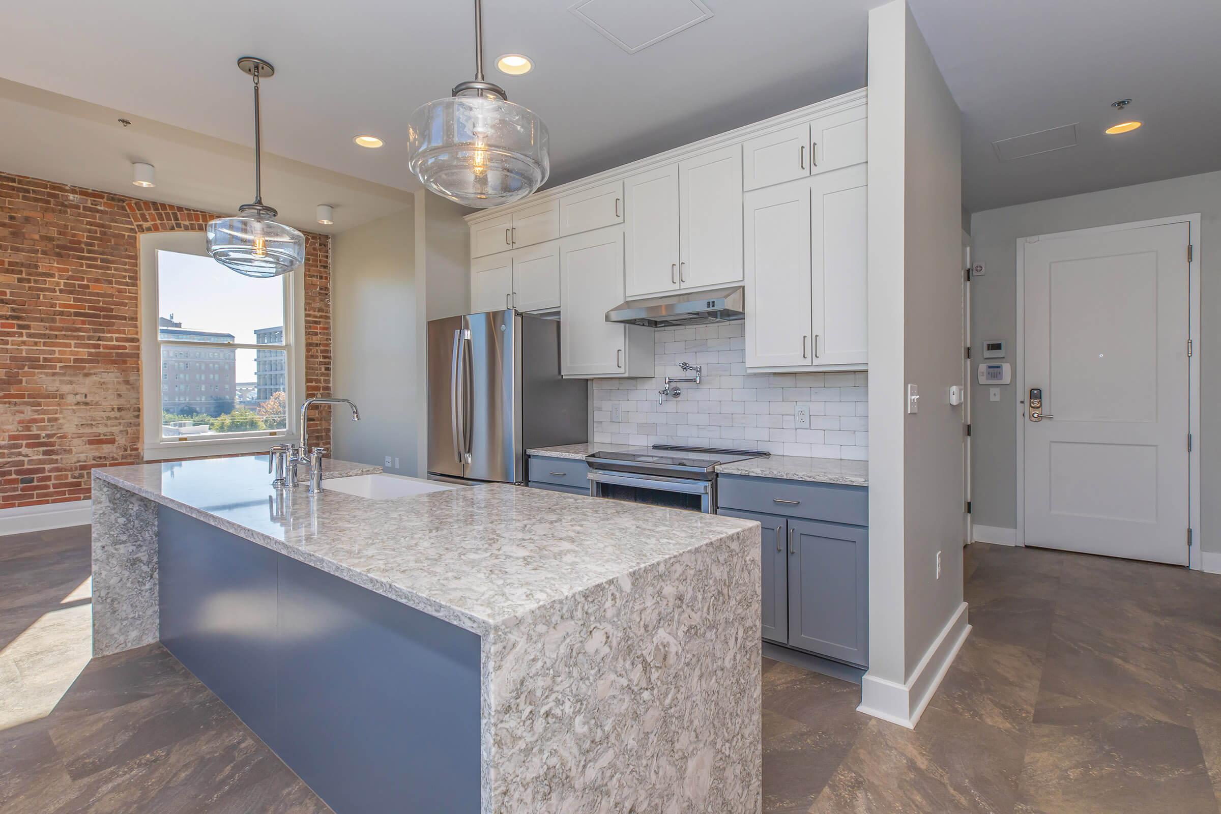 A modern kitchen with a large gray island, white cabinets, and stainless steel appliances. Features include a sleek countertop, a brick accent wall, and large windows allowing natural light. The floor is tiled, and there is a door leading to another room. Stylish pendant lights hang above the island.