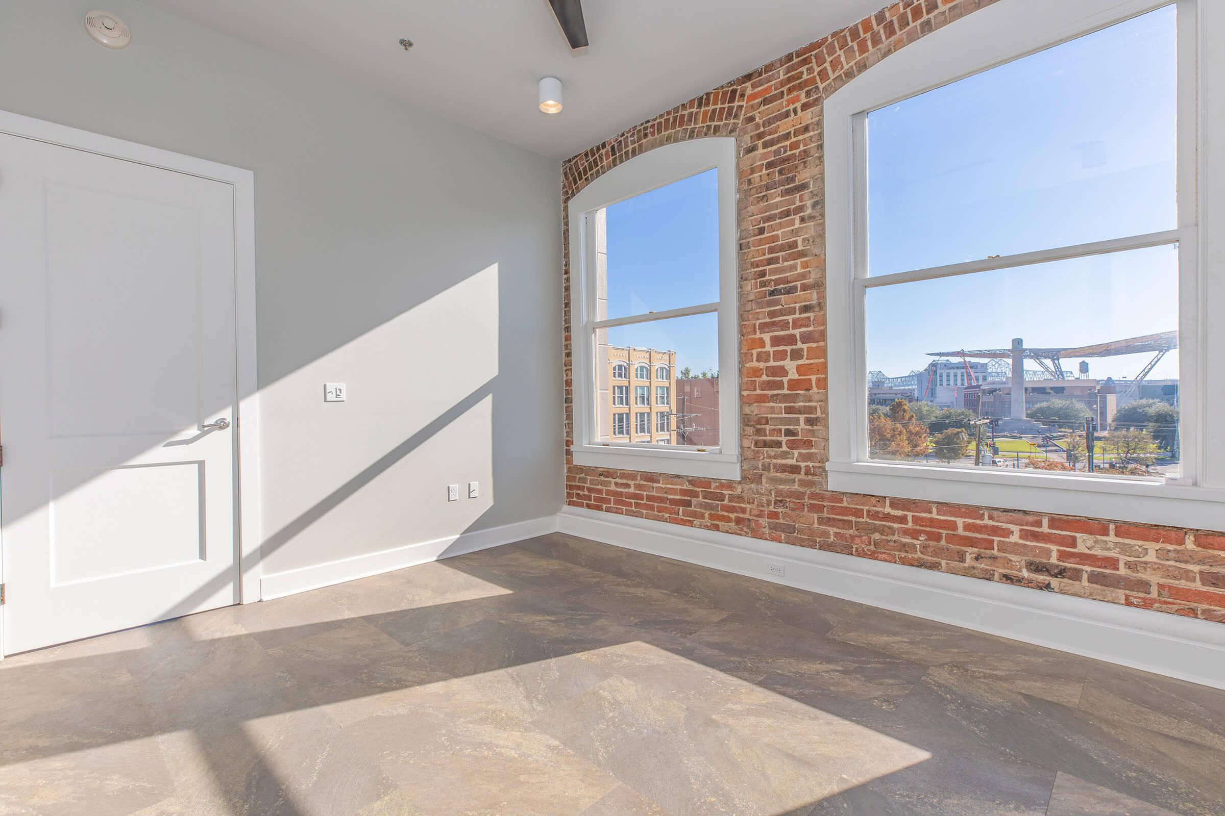 Bright interior of a modern room featuring large windows with a view of a stadium. Exposed brick walls add character to the space, complemented by neutral-colored walls and flooring. Natural light floods the room, creating a warm and inviting atmosphere.